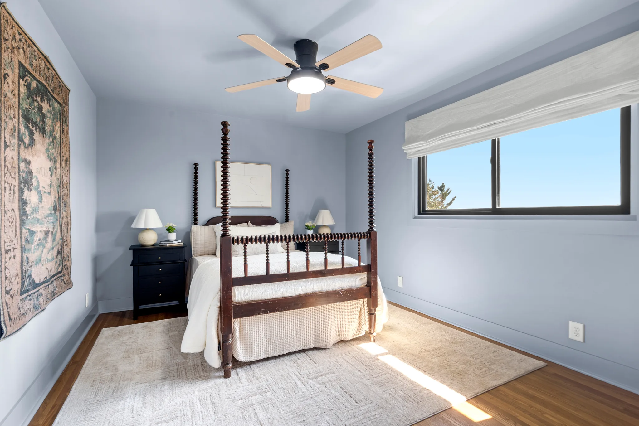 Bedroom with light gray walls, hardwood floors, a dark four-poster bed, black nightstands, ceiling fan and a wide window with roller shade