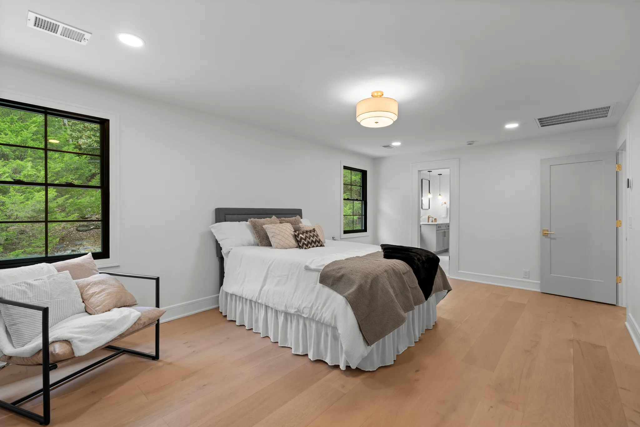 Bedroom with white walls, wide oak plank flooring, a gray upholstered bed, black grid window overlooking trees and an open door to a bathroom vanity