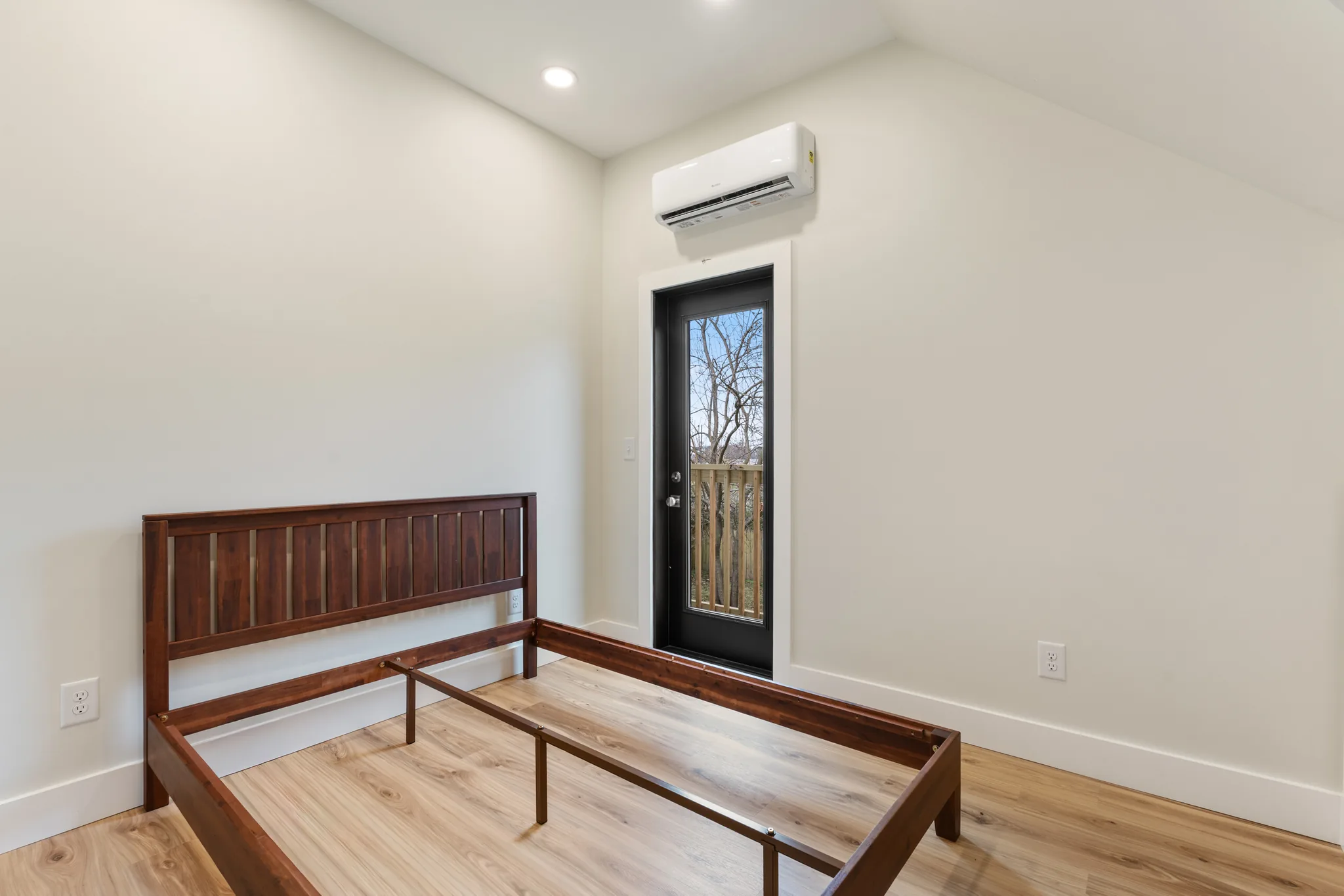 Finished bedroom with white walls, light wood floors, a wall-mounted mini-split unit and a glass door leading to a fenced yard