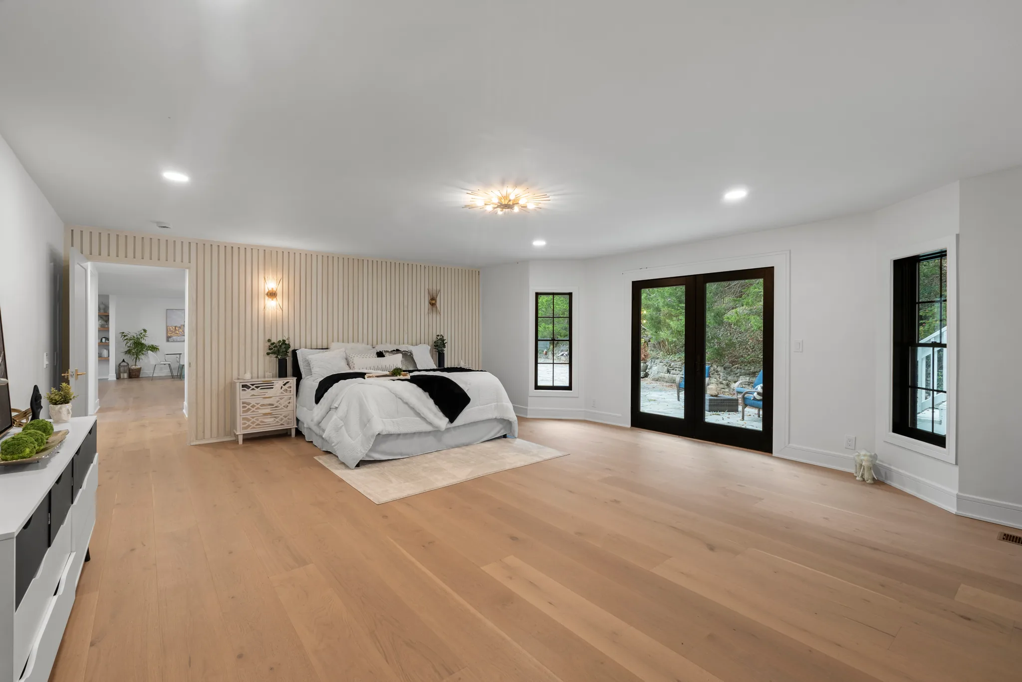 Large bedroom with vertical wood slat accent wall behind the bed, wide oak plank floors, black French doors and black grid windows
