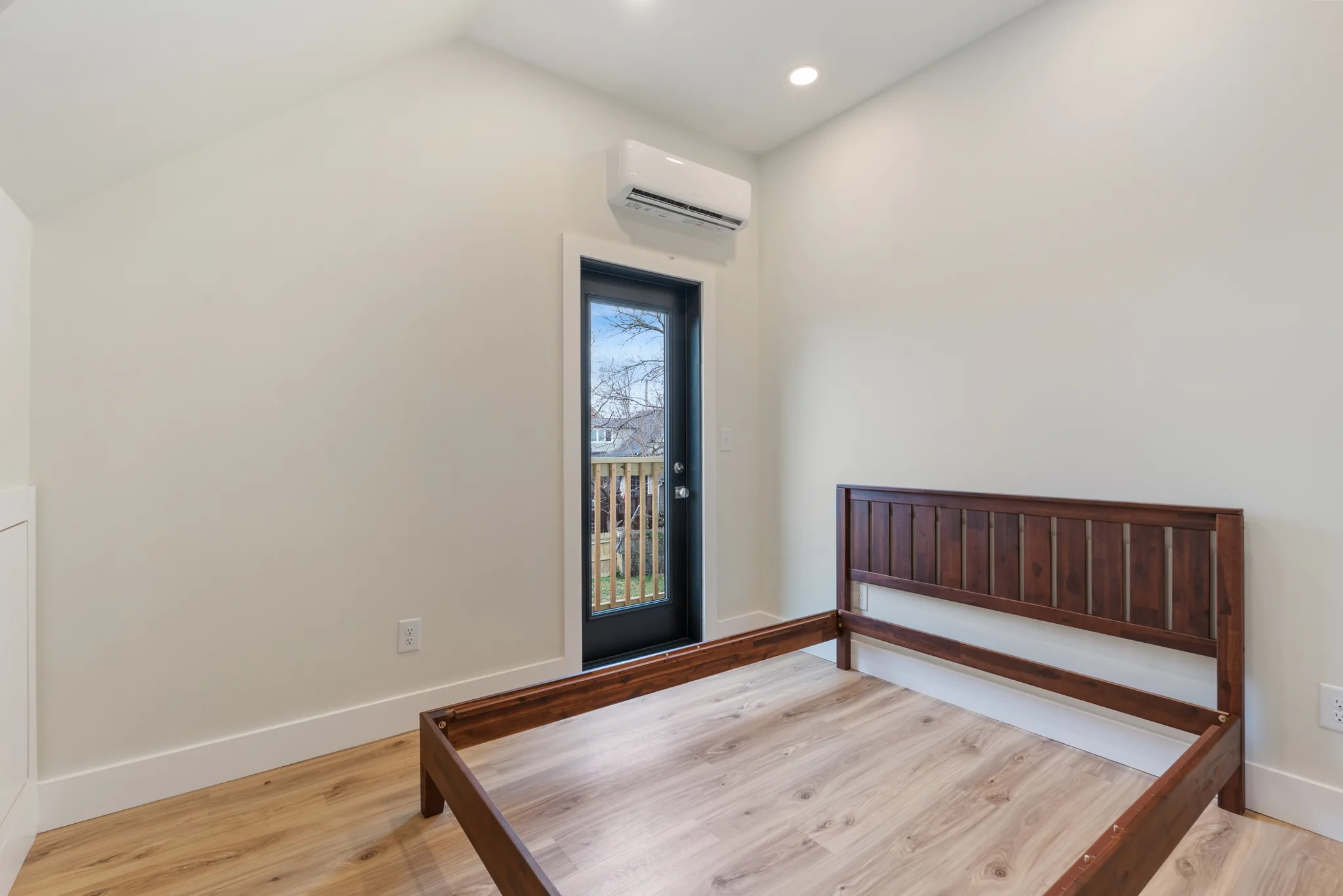 Finished bedroom with sloped white ceiling, wall-mounted mini-split, wood bed frame and a glass door opening to a balcony