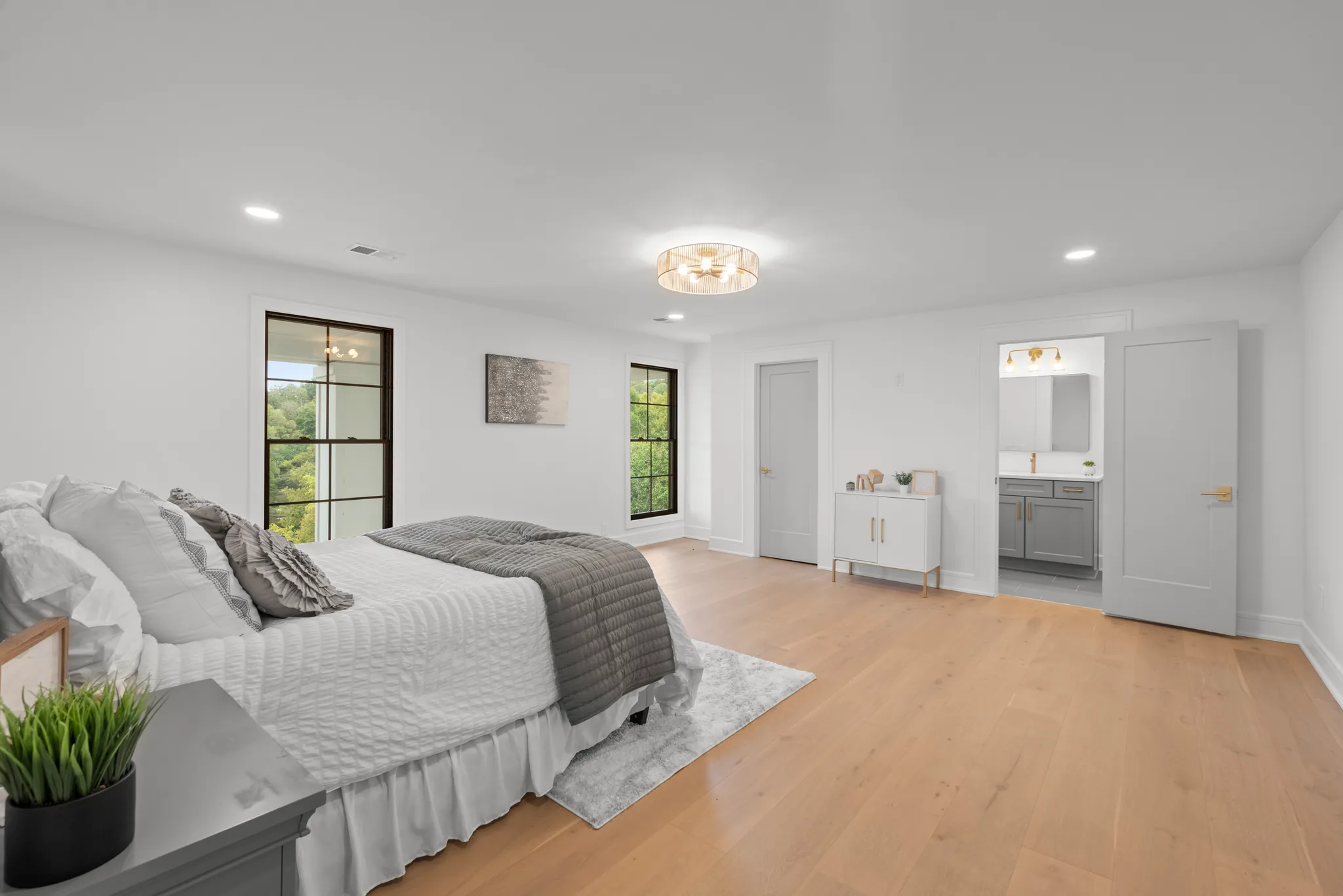 Bedroom with white walls, wide-plank oak floors, black grid windows and an open door showing a gray vanity in the adjoining bath