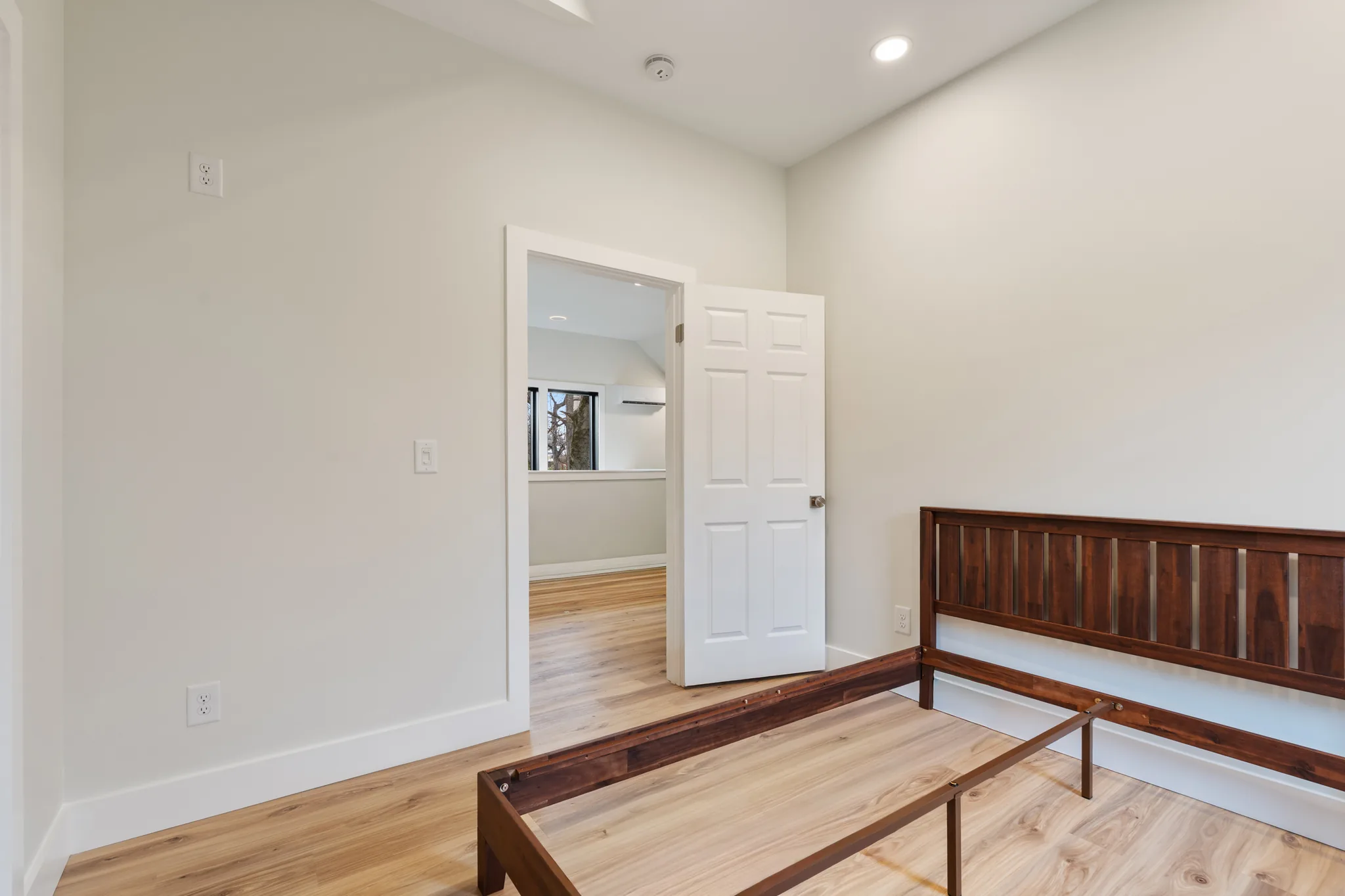 Finished bedroom with off-white walls, light wide-plank wood floors and a dark wood bed frame beside an open six-panel door