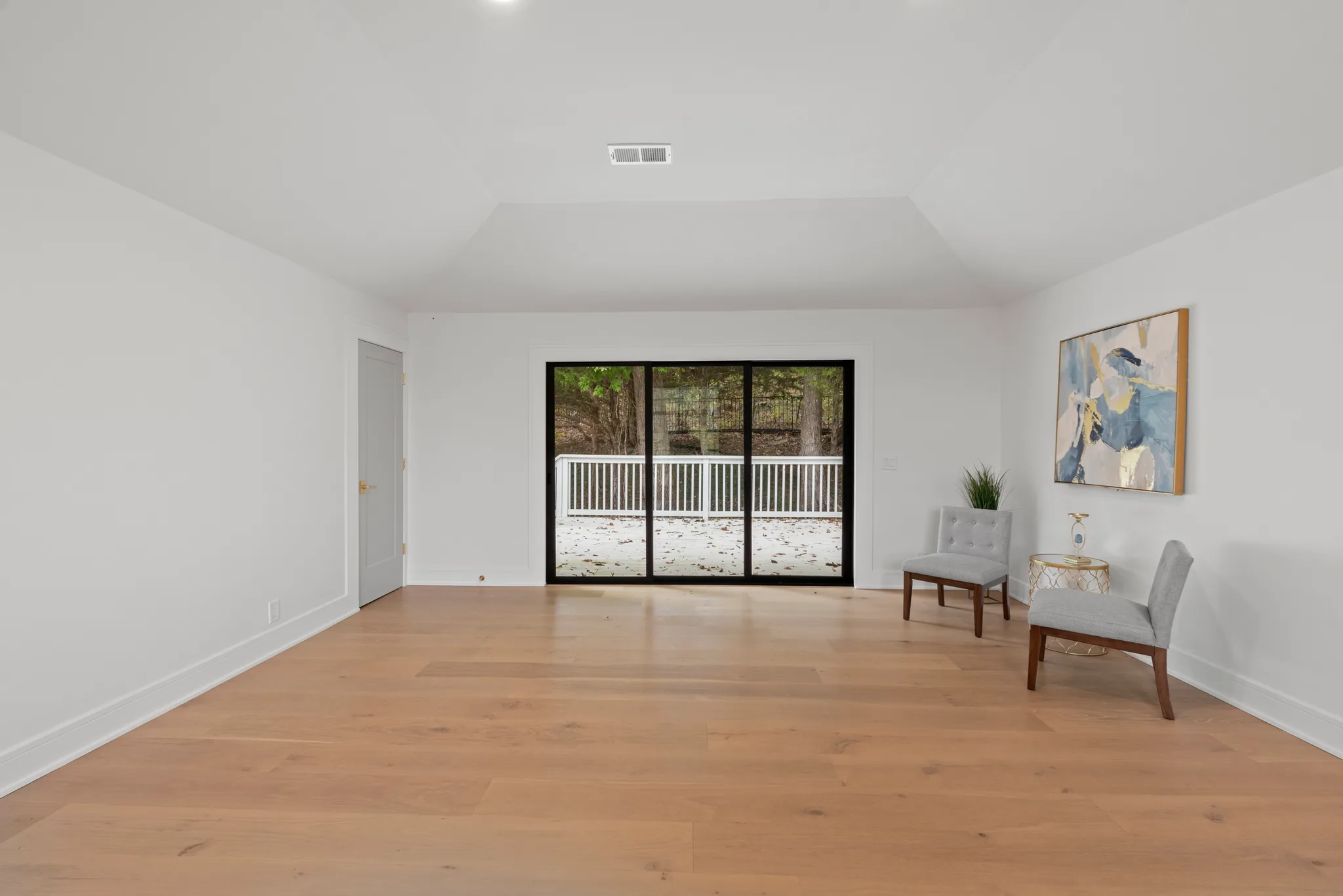Open bonus room with white walls, wide oak flooring and a black three-panel sliding glass door leading to a white-railed deck and woods