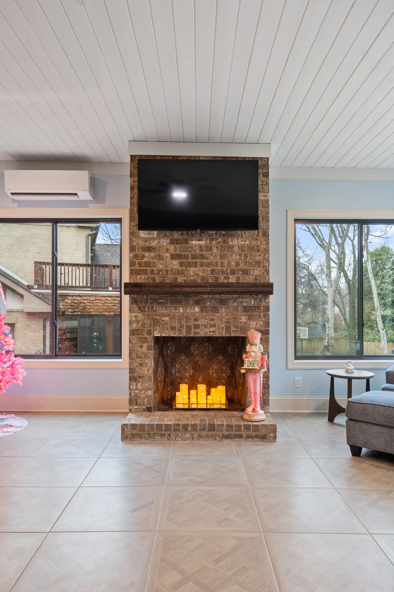 Interior sitting room with a floor-to-ceiling brick fireplace, wood mantel, mounted TV, white plank ceiling, large tile floor and windows on both sides