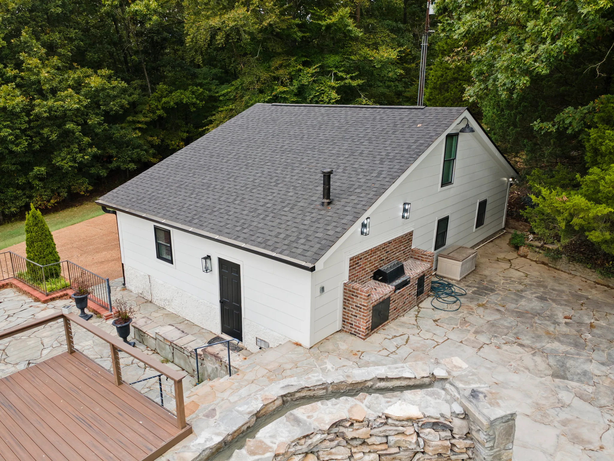Aerial view of a white lap-sided outbuilding with dark shingle roof, brick built-in grill alcove and wide flagstone patio beside a wood deck