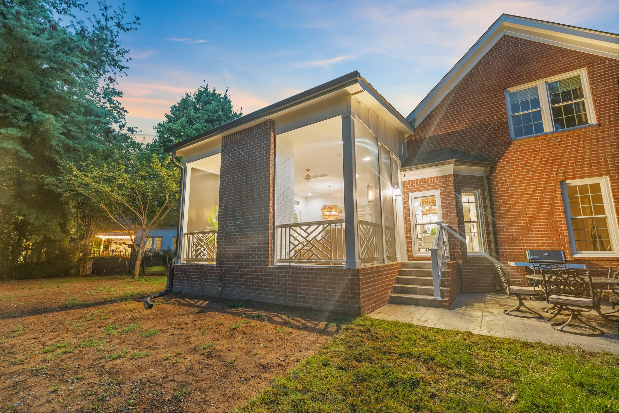 Screened porch addition with brick piers, Chippendale-style railing and lit interior attached to a two-story brick home at dusk