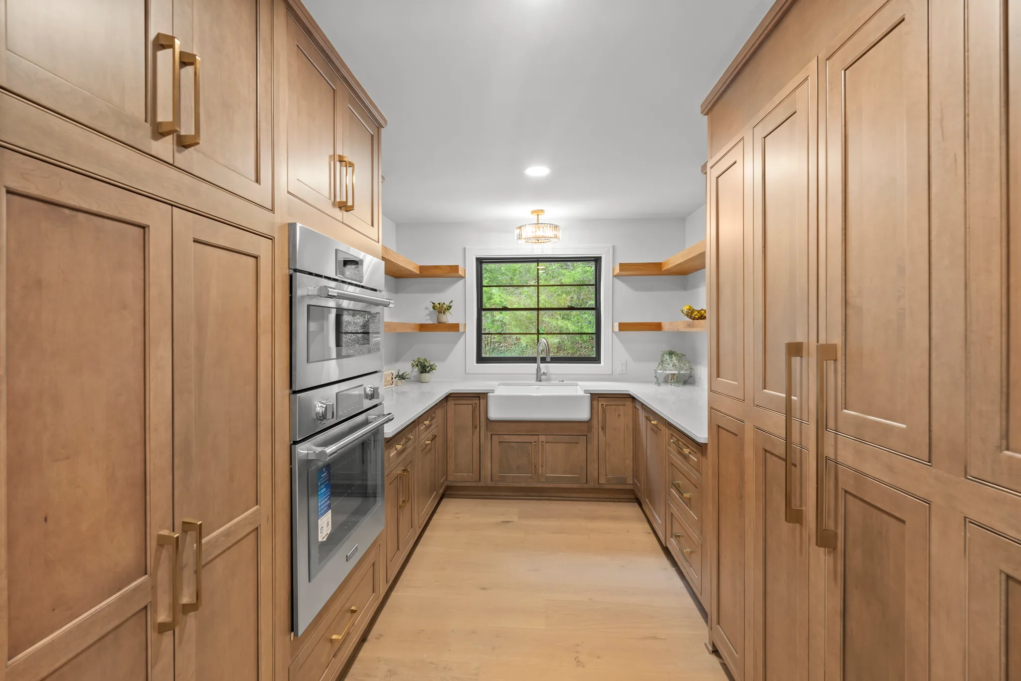 Galley-style butler pantry with stained wood cabinets, brass pulls, stainless double wall ovens, farmhouse sink under a black grid window and floating wood shelves