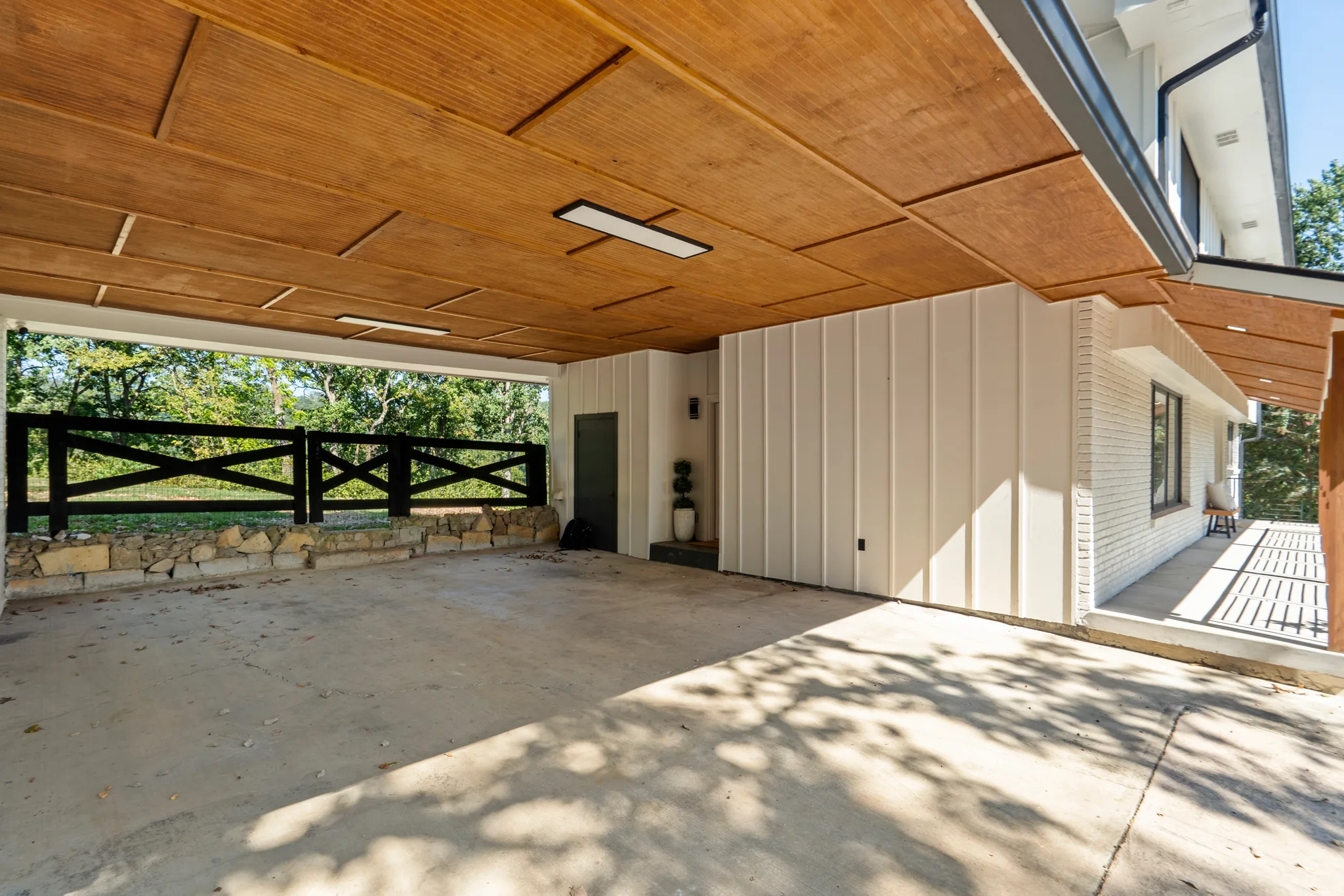 Covered carport with a stained tongue-and-groove wood ceiling, white board-and-batten walls, concrete slab, and a black fence over a stone retaining wall beyond
