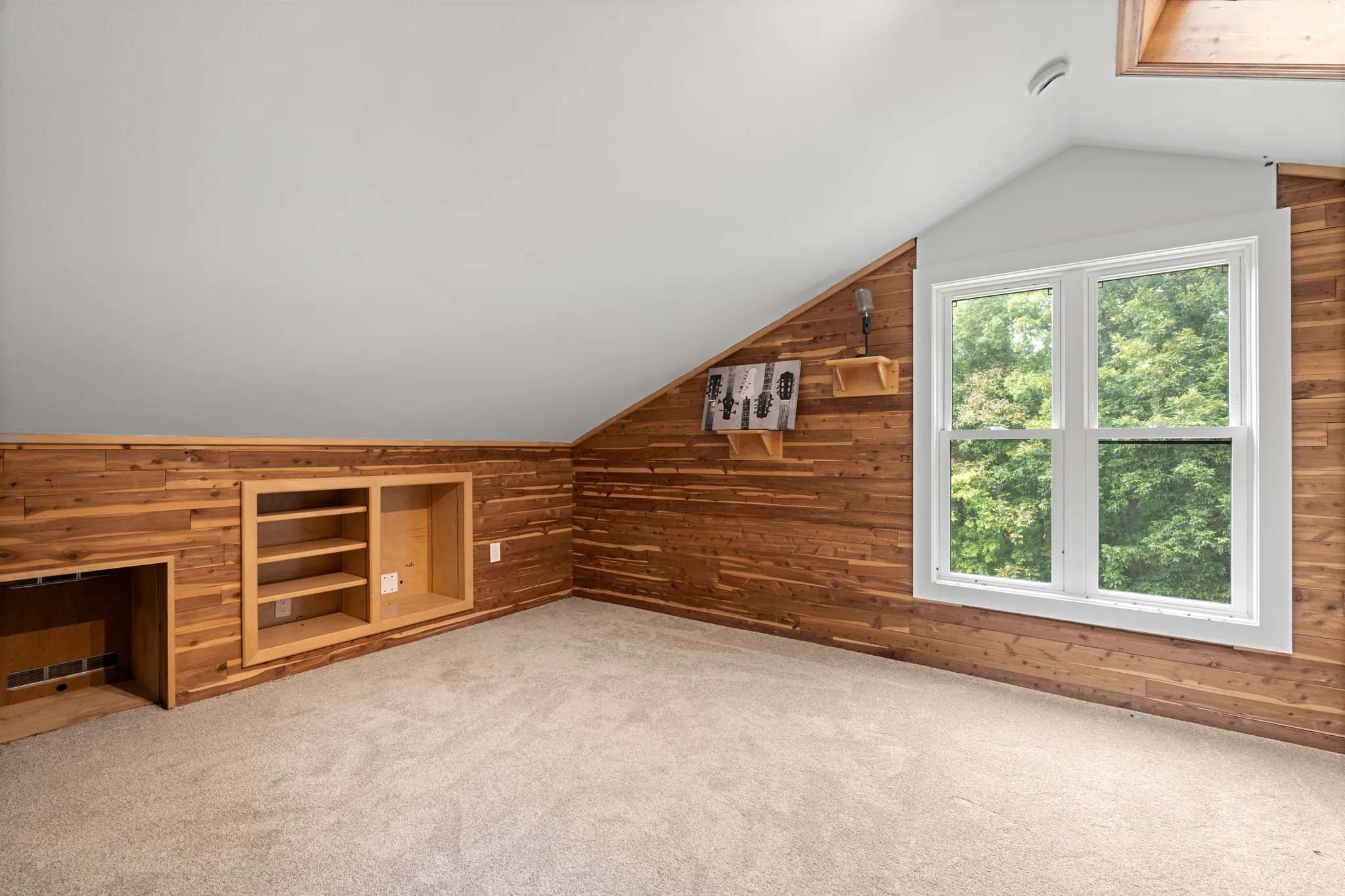 Empty upstairs bonus room with cedar plank walls, sloped white ceiling, carpeted floor, built-in shelving and a white double-hung window facing trees