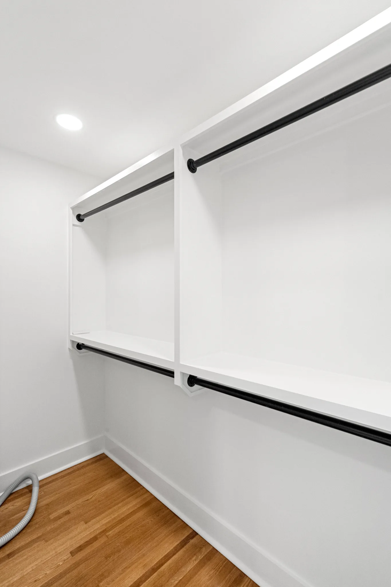 Empty walk-in closet with white shelves, black metal hanging rods on two levels, white walls and oak flooring