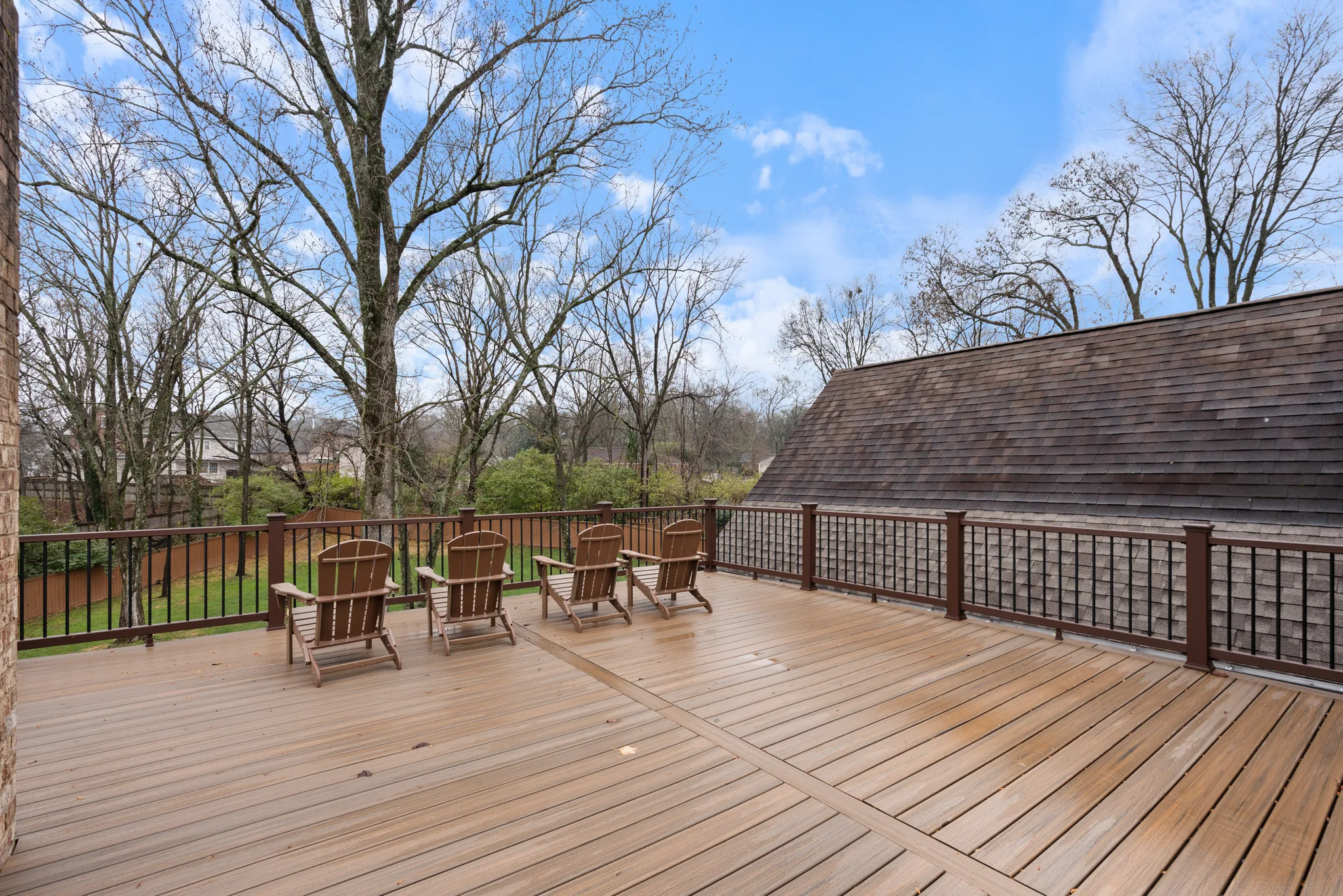 Wide composite deck with tan boards, dark railings and black balusters set beside a shingled roof slope, with bare trees beyond