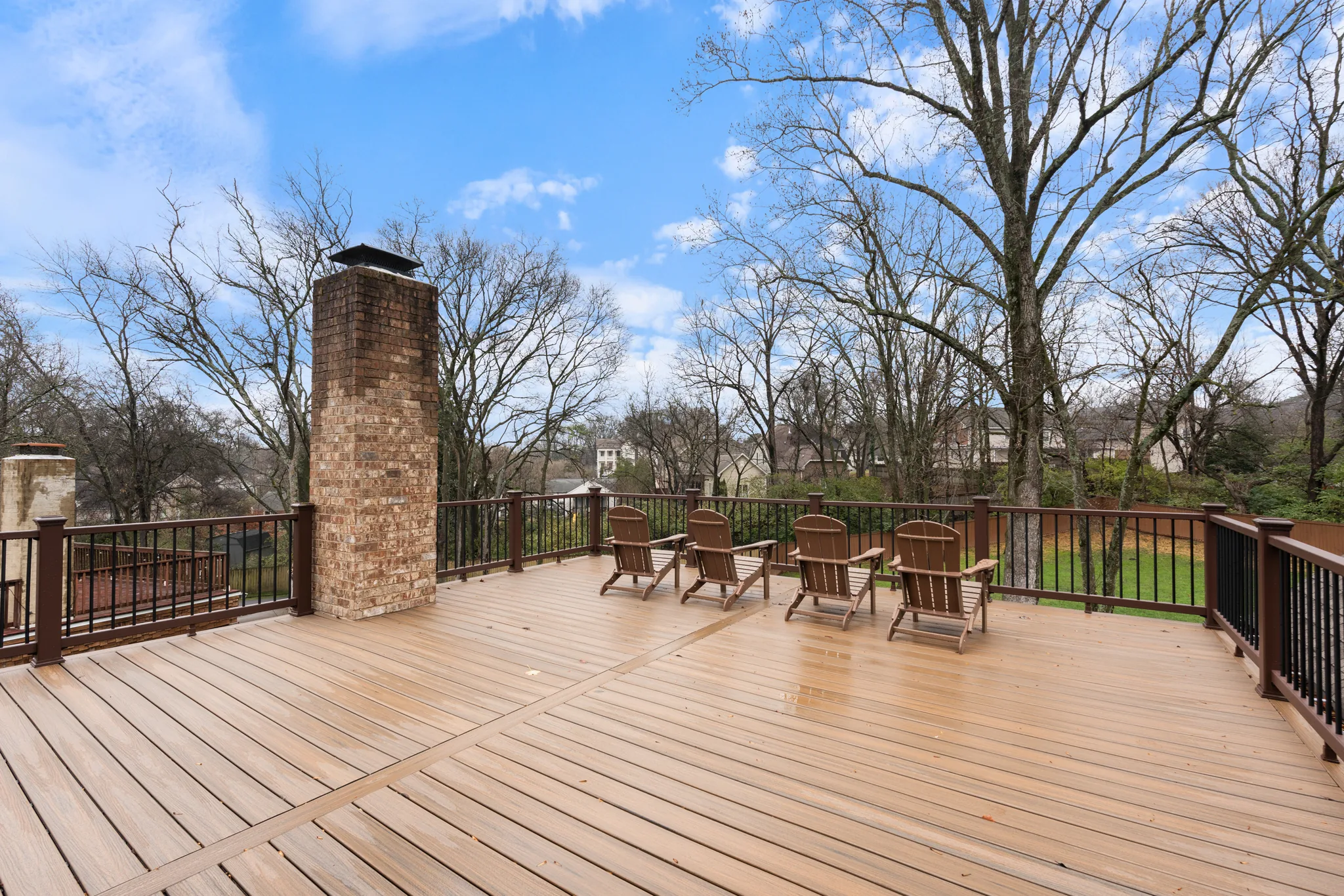 Wide composite deck with tan boards, brick chimney stack and black metal railings, holding four wood Adirondack chairs above a wooded yard