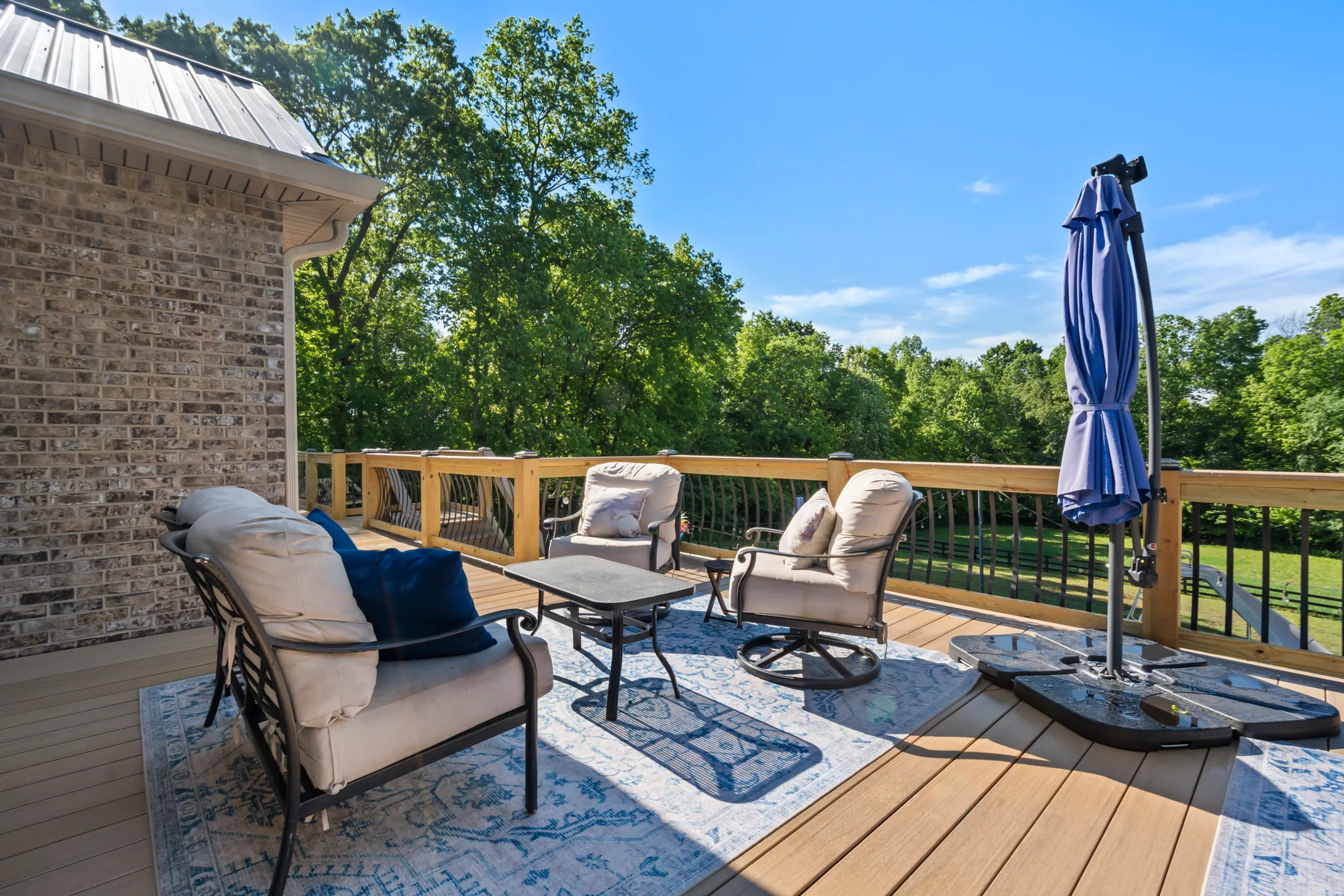 Composite deck beside a brick home with cushioned patio chairs, a rug, a folded blue cantilever umbrella and wood railing facing trees
