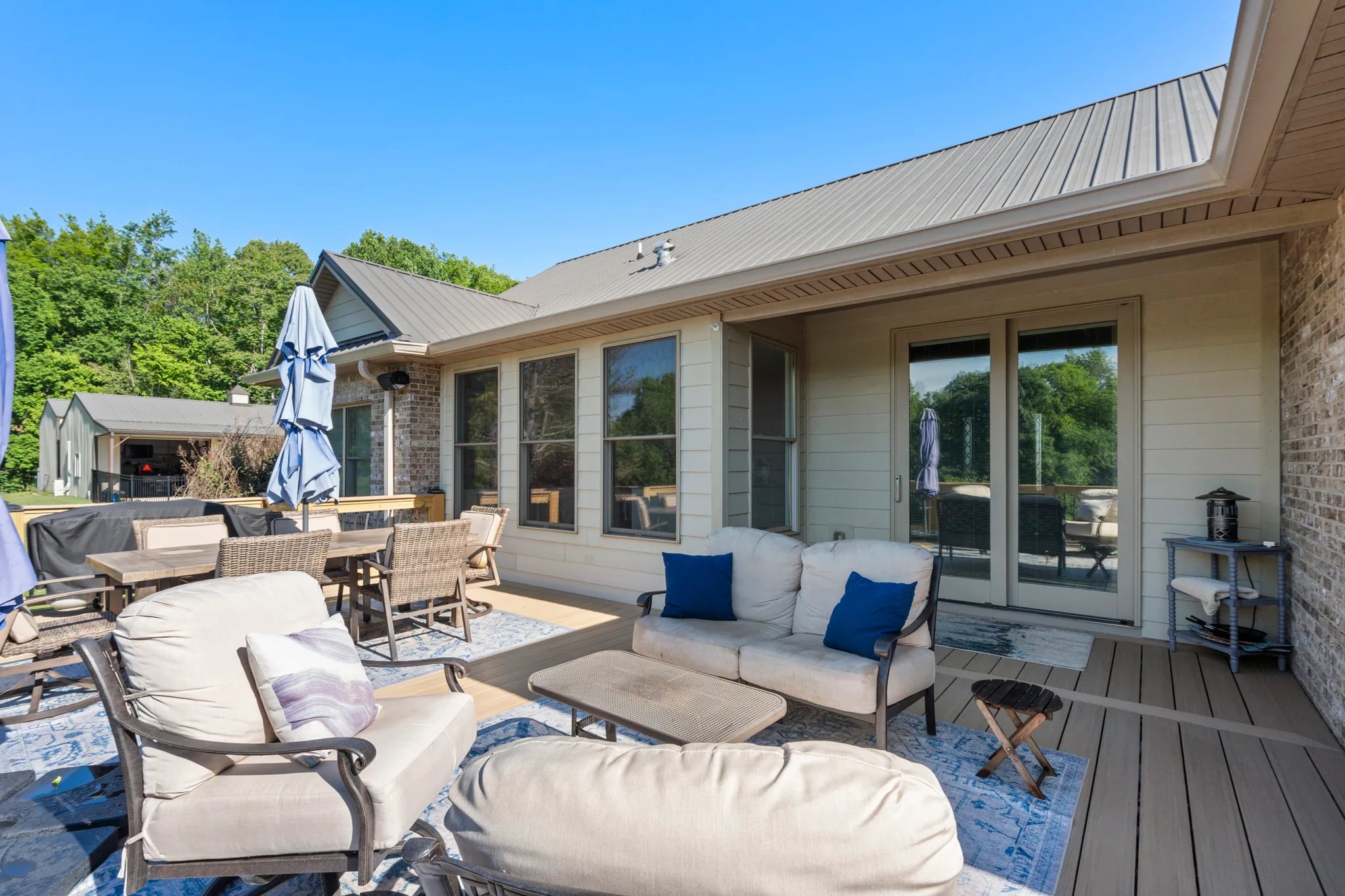 Composite deck with cushioned lounge chairs, patterned outdoor rugs and a closed blue umbrella beside a home with siding and metal roof