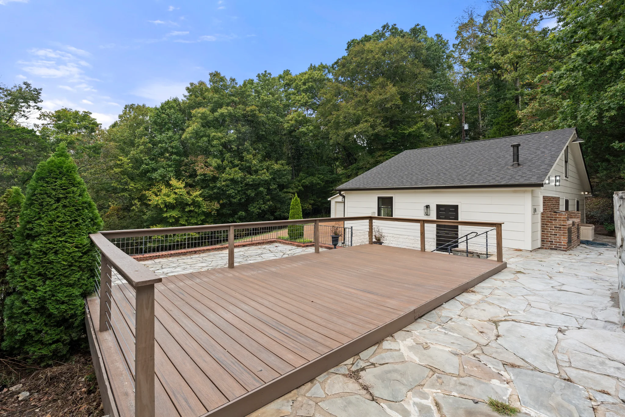 Low composite deck with cable railing and stained wood top rail next to an irregular flagstone patio behind a white gabled house in the woods