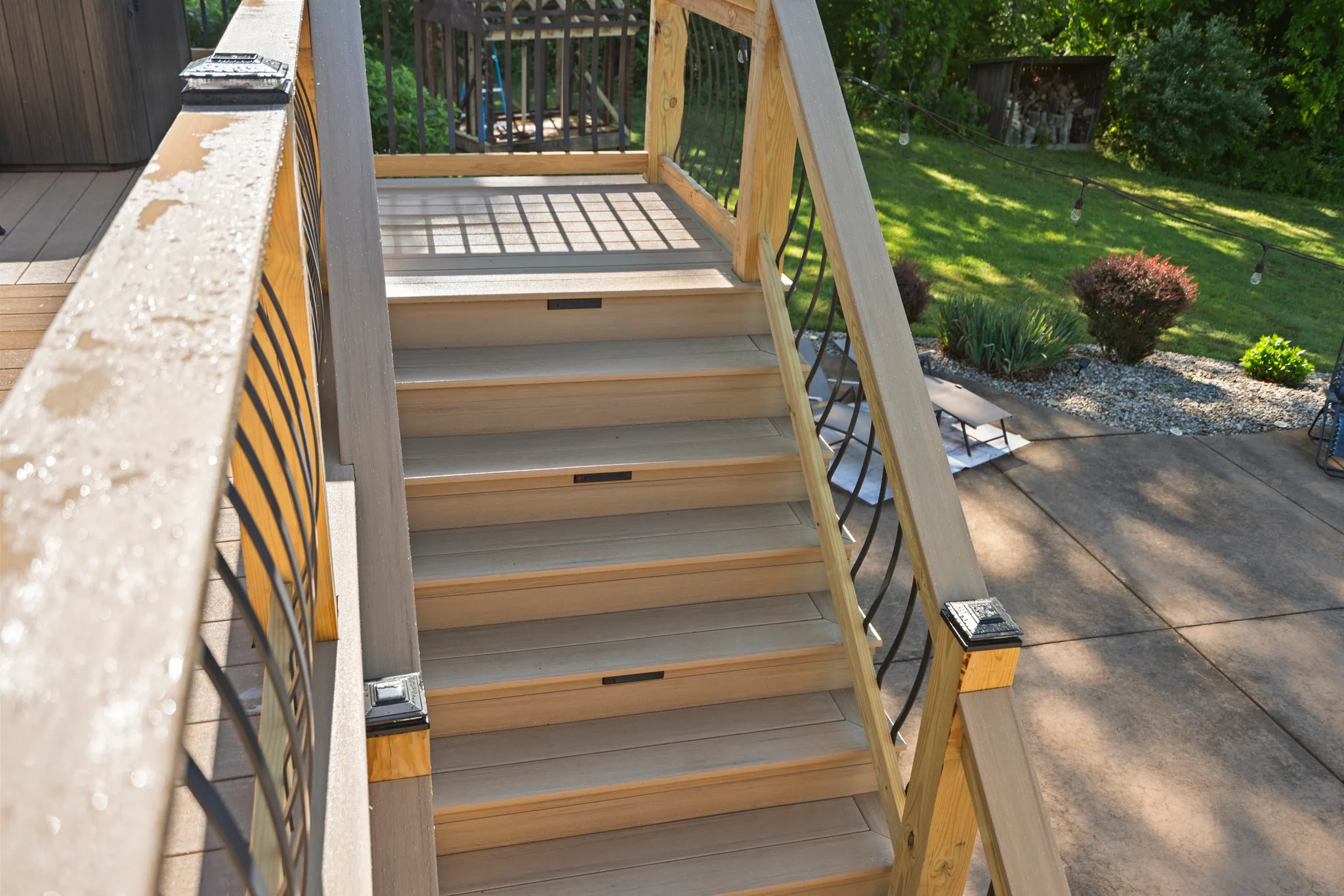 Composite deck stairs with gray treads, wood posts, black post caps and curved metal balusters descending to a concrete patio beside a lawn