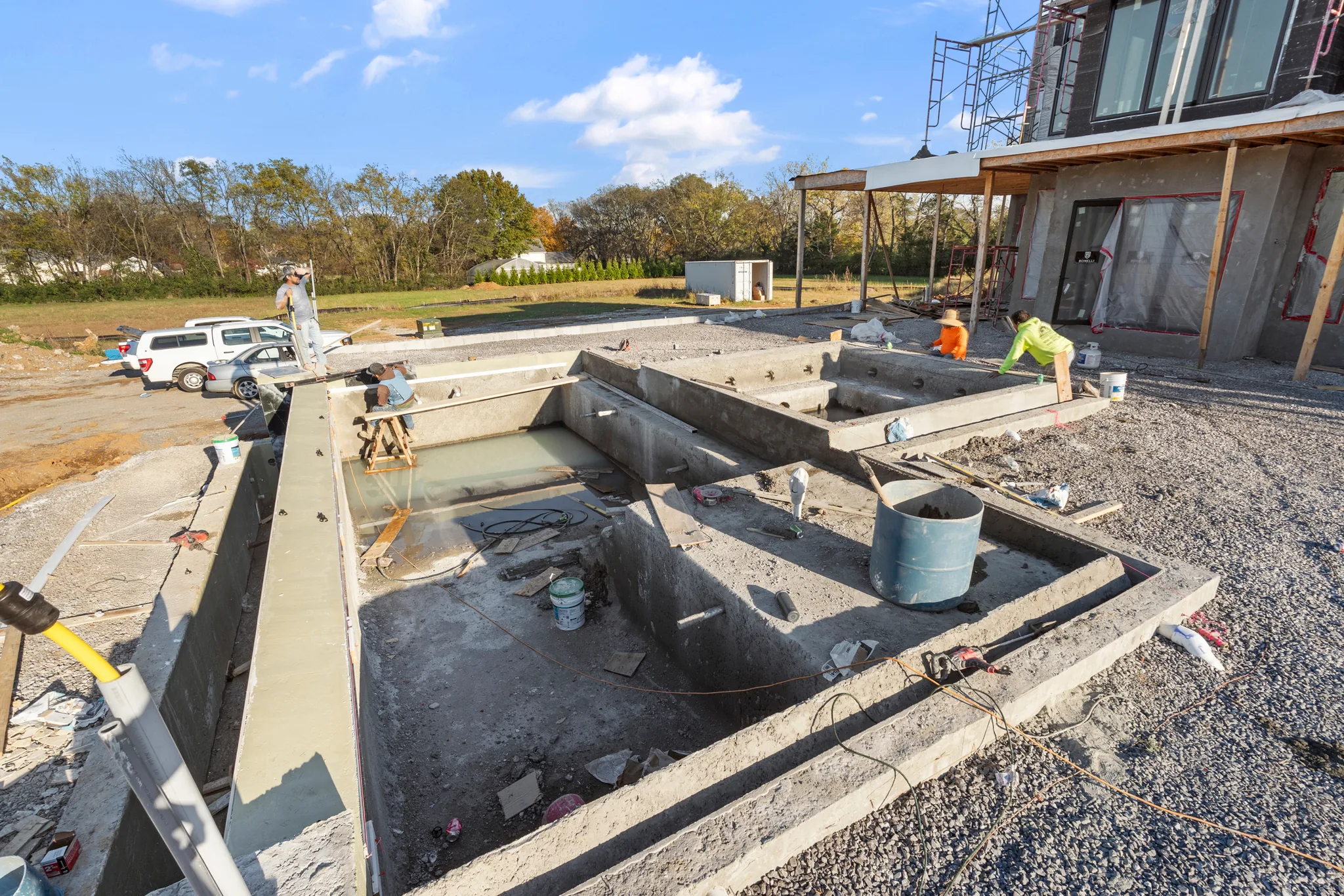 Poured concrete pool and spa shells under construction on a gravel yard beside a two-story modern home wrapped in sheathing with scaffolding