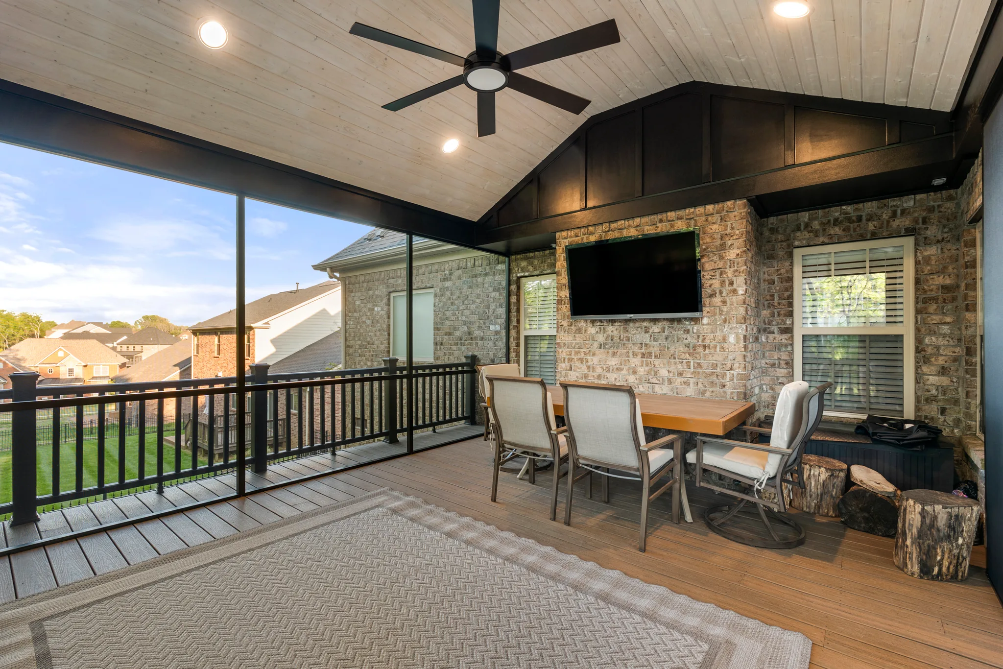 Covered deck with vaulted whitewashed wood ceiling, black fan, TV mounted on brick, dining set and black metal railing overlooking a lawn