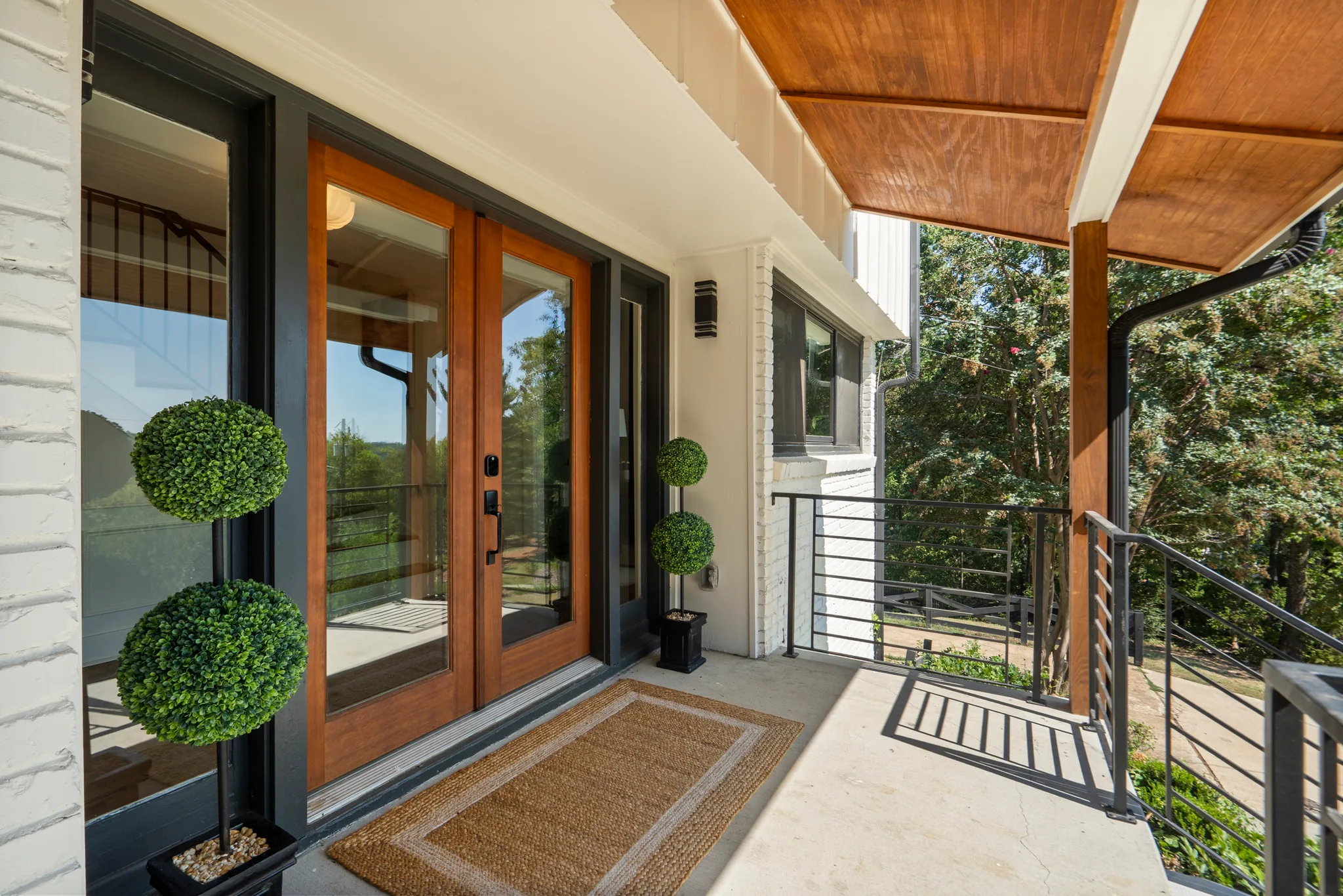 Covered front porch with stained wood plank ceiling, glass entry door in wood frame, painted brick walls and horizontal cable railing