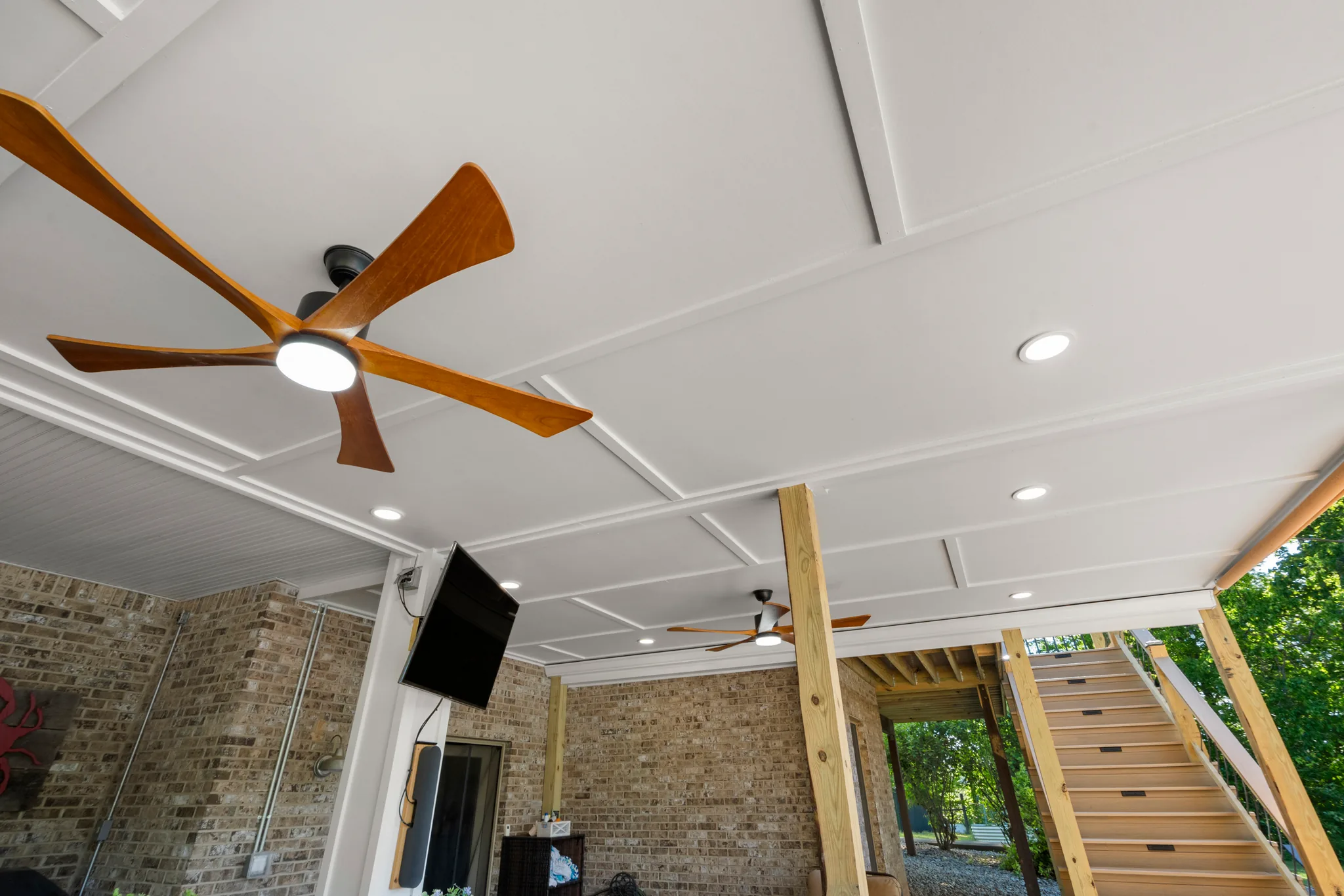 Covered lower patio with white paneled ceiling, two wood-blade ceiling fans, recessed lights, brick walls, mounted TV and wood stairs