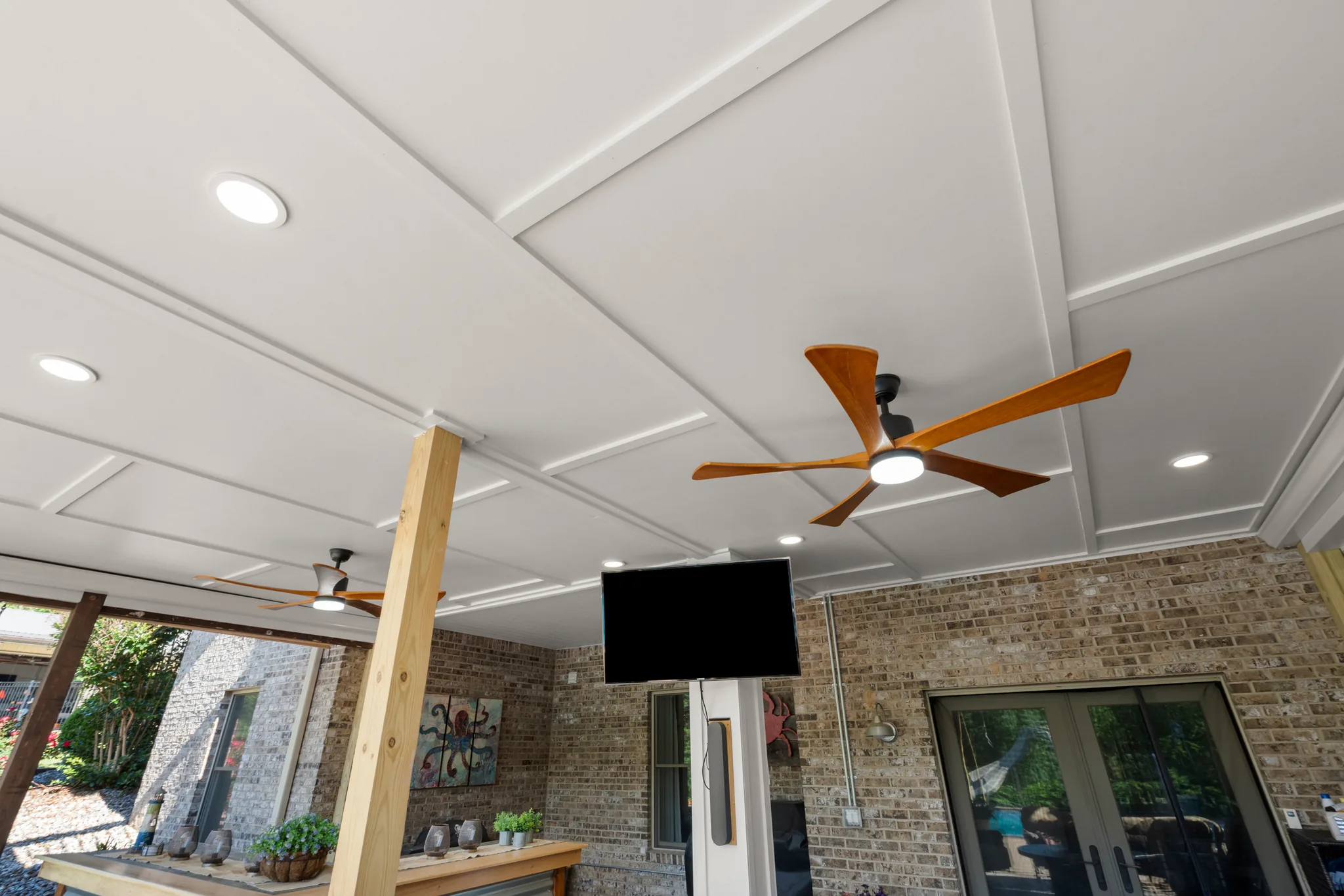 Covered outdoor patio with a white coffered ceiling, recessed lights, two wood-blade fans, mounted television, brick walls and glass doors