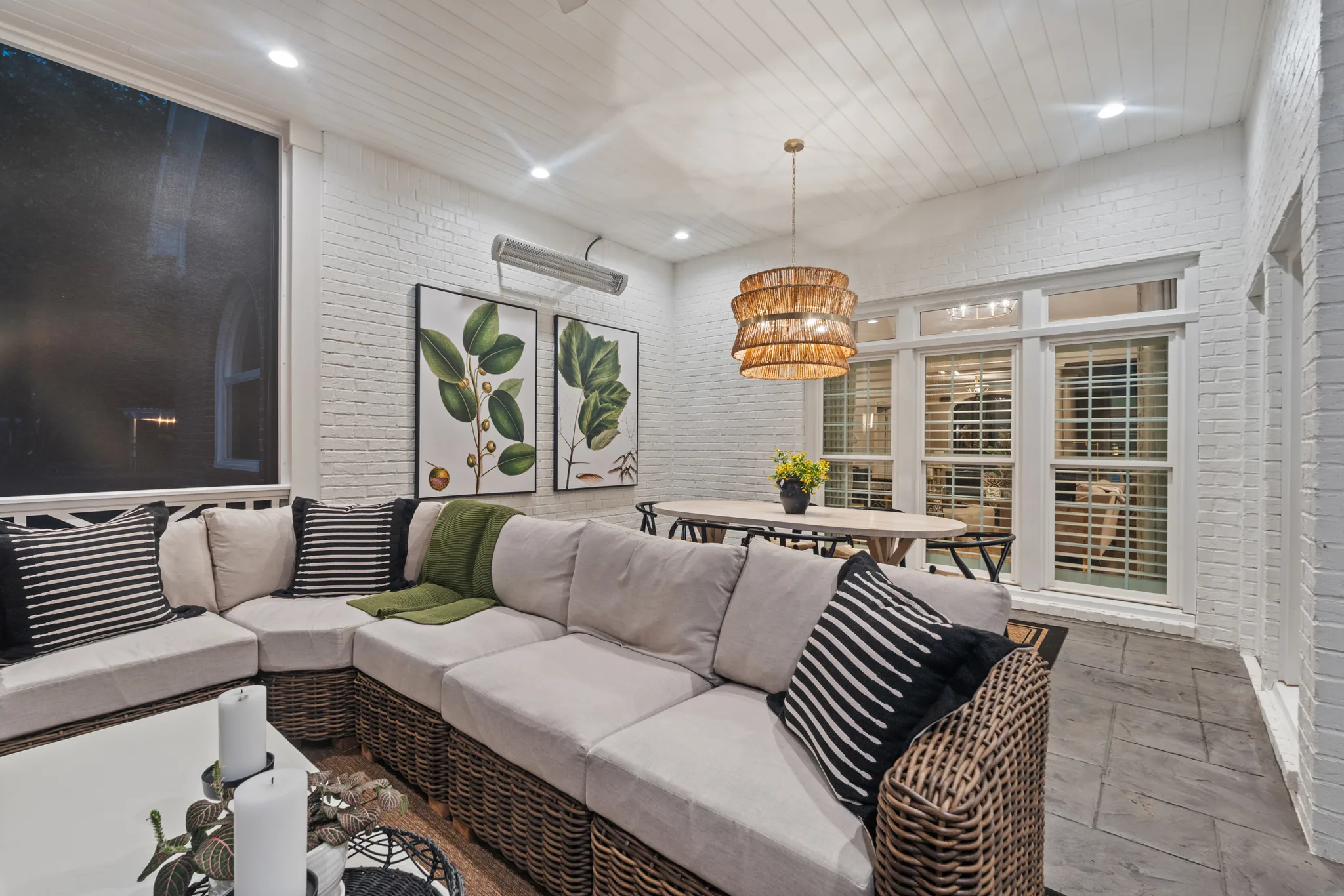 Covered porch with painted white brick walls, white plank ceiling, woven sectional, botanical prints, rattan pendant over a dining table, and tile floor