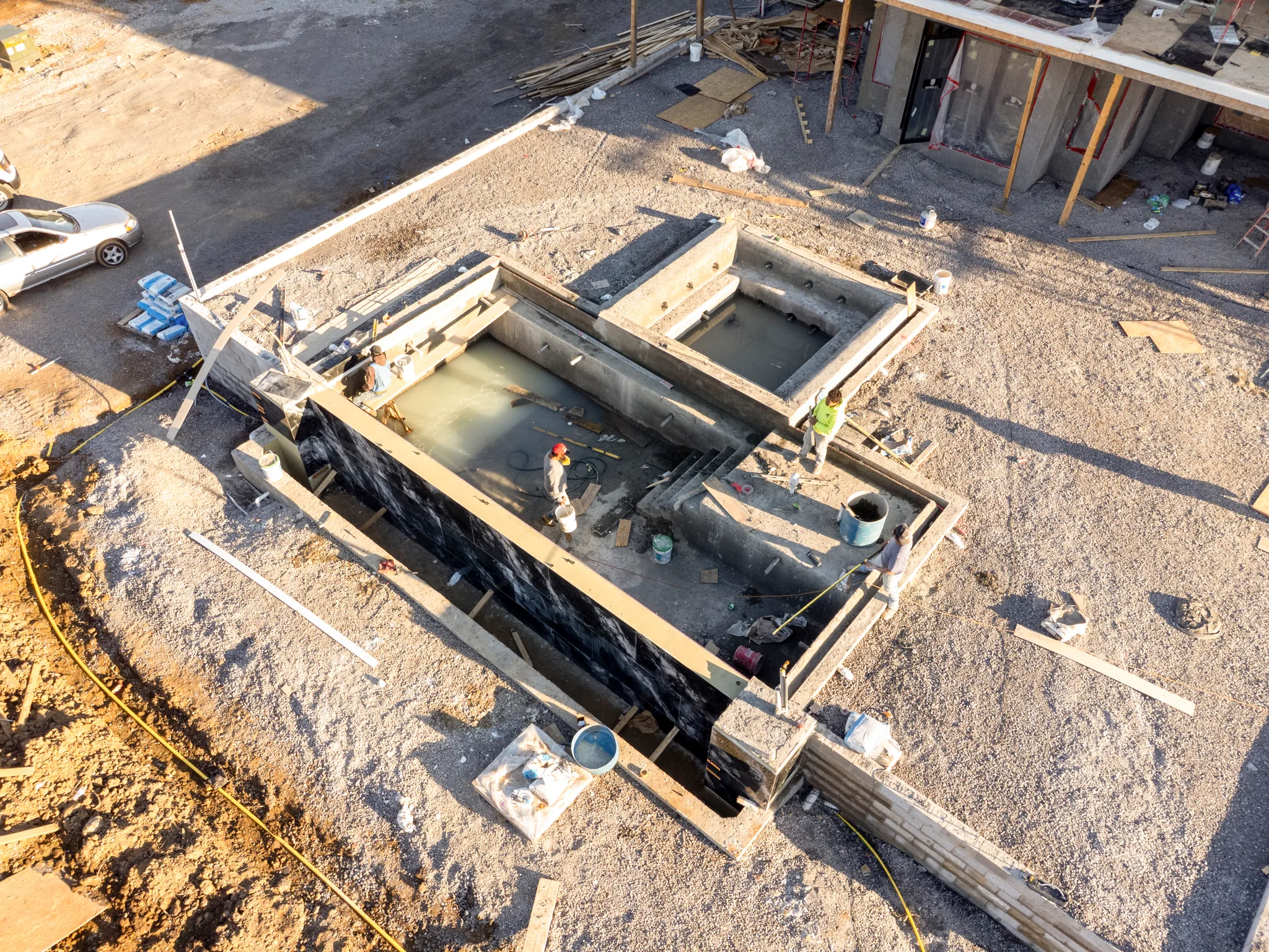 Aerial view of a concrete pool shell under construction with black waterproofing membrane on one wall and workers inside the excavation on a gravel site