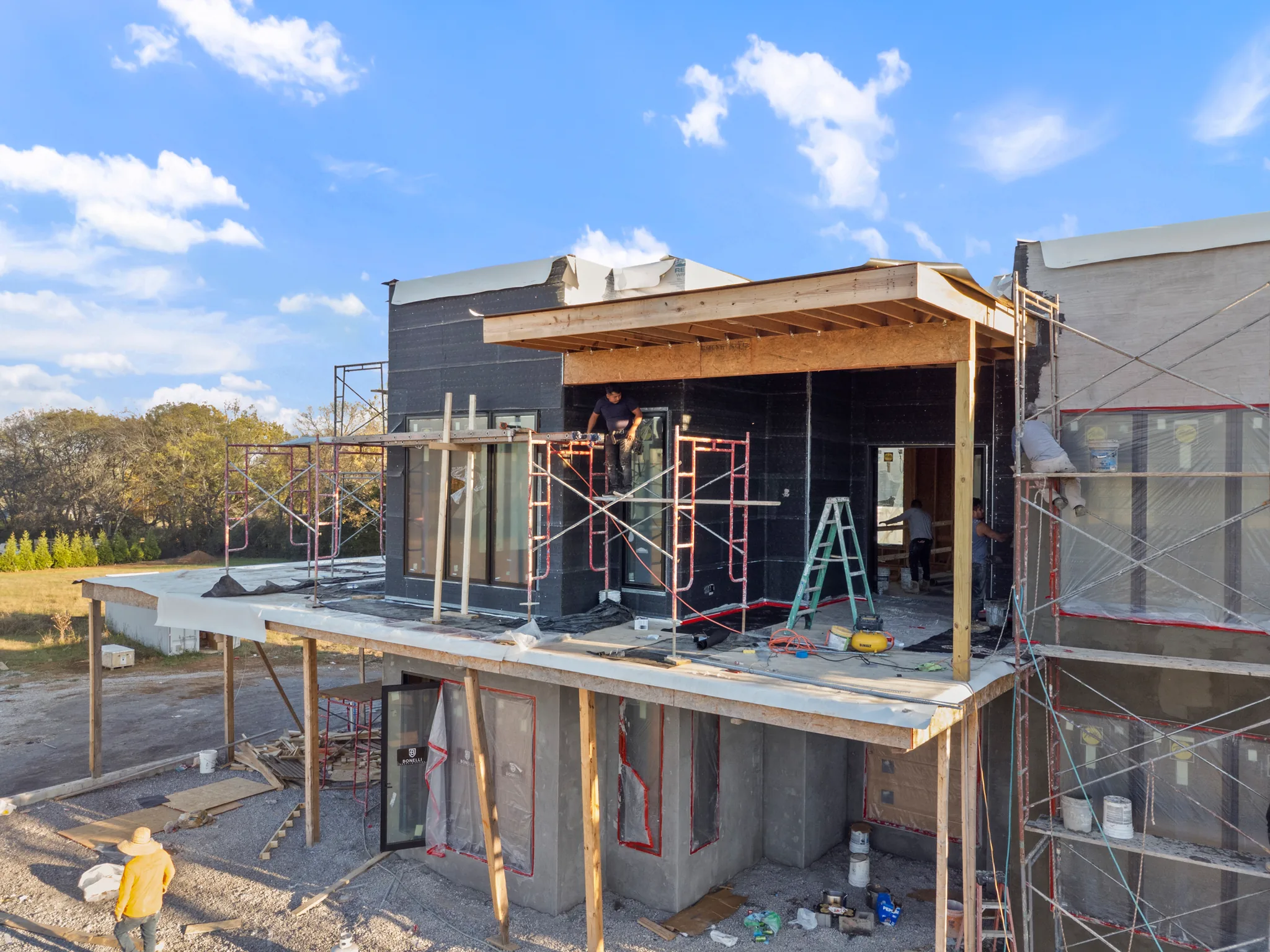 Workers on scaffolding installing glass doors and windows on a modern home under construction wrapped in black weather barrier with exposed wood framing