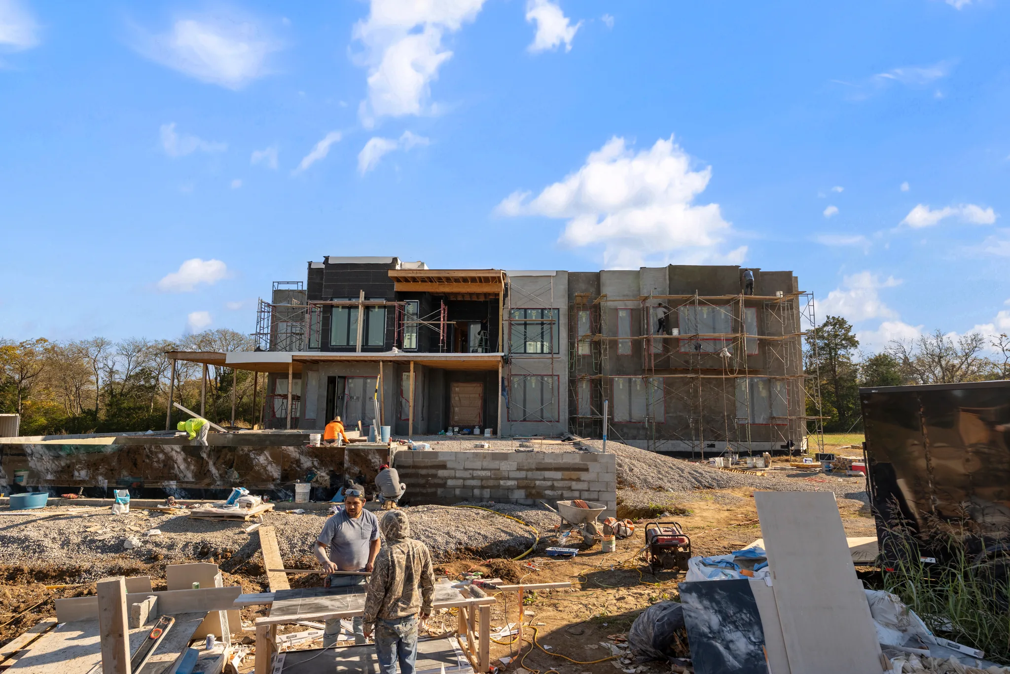 Modern two-story home under construction with scaffolding and workers setting stone veneer on a retaining wall in the foreground