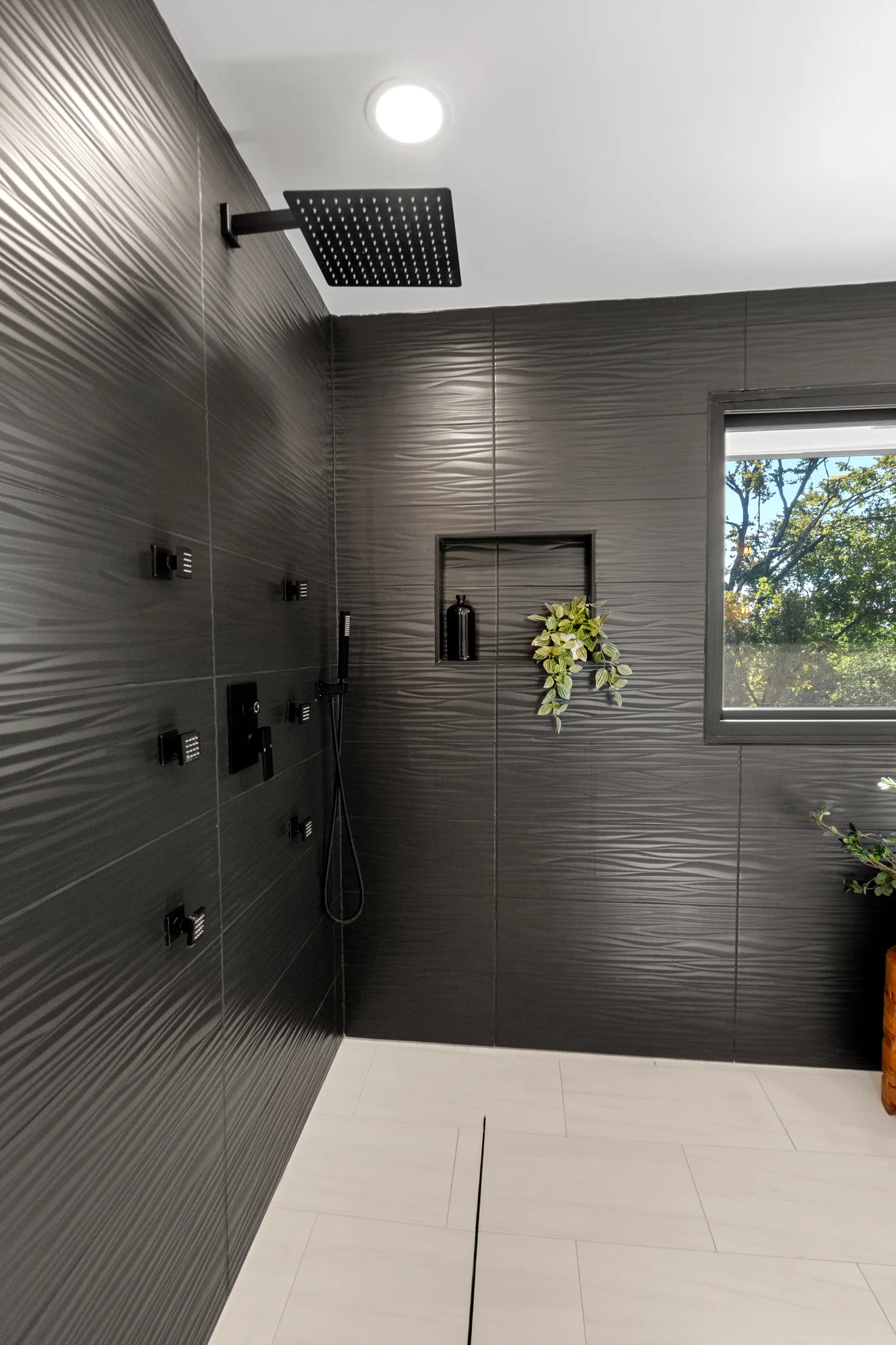 Walk-in curbless shower lined with textured charcoal tile, matte black rain head, body sprays, recessed niche, linear drain and window