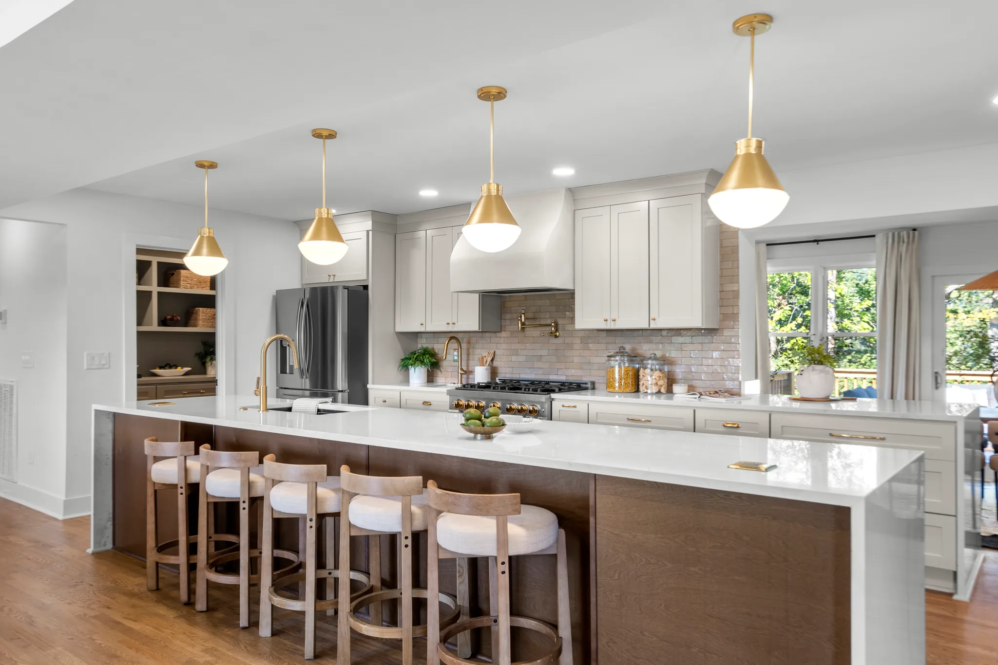 Kitchen with a long dark wood island topped in white quartz, five light wood stools, brass dome pendants and pale gray cabinets