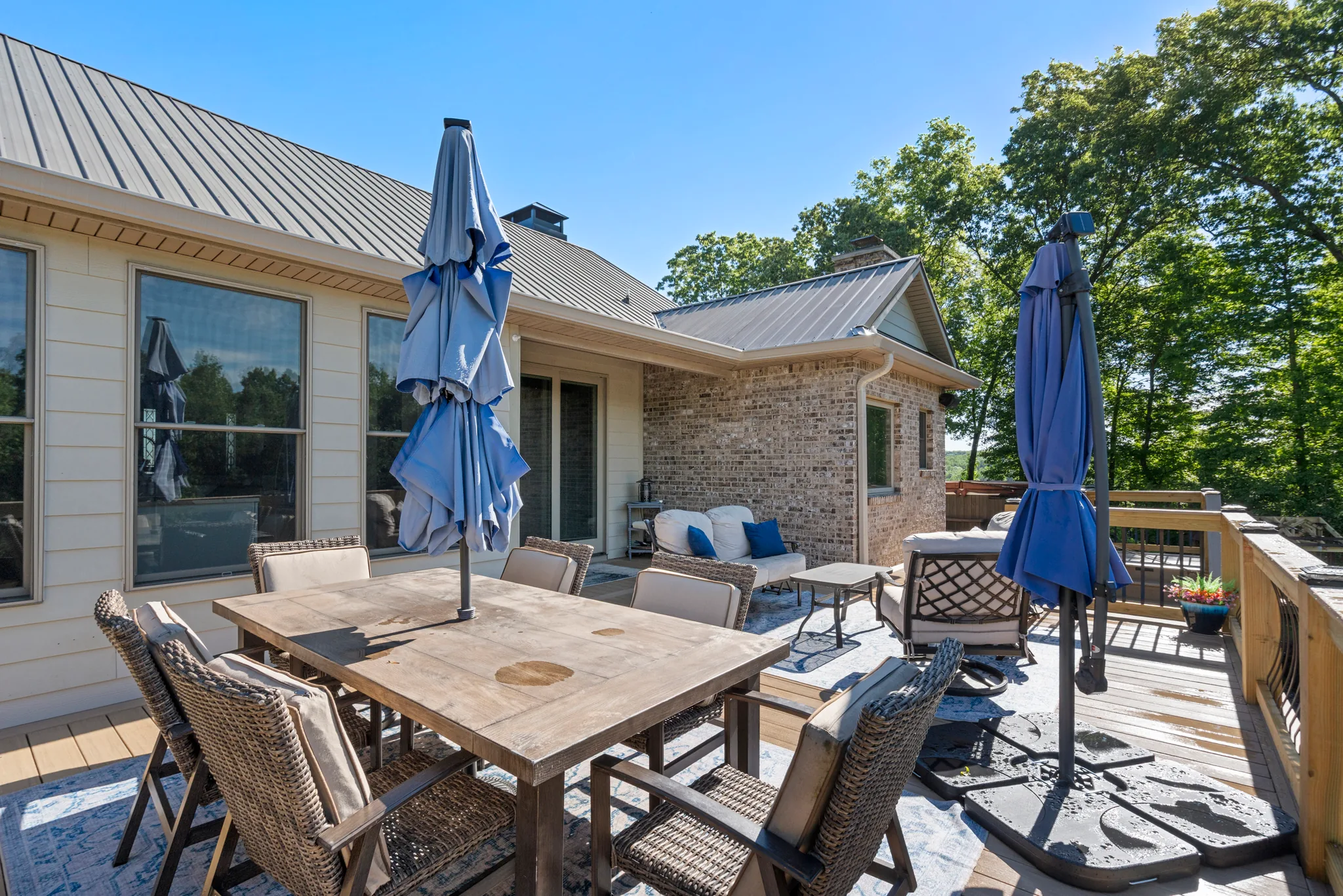 Wide deck with a wood dining table, wicker chairs and two closed blue umbrellas beside a brick and lap-sided home with a metal roof