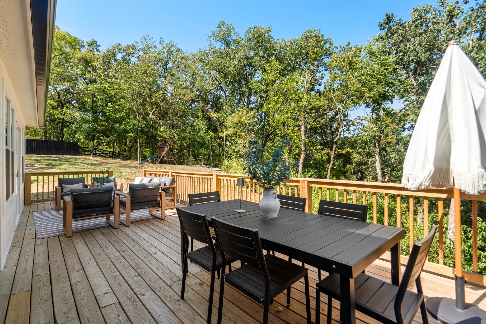 Wood deck with a black outdoor dining table and chairs, white patio umbrella, and lounge seating beside a wood railing overlooking trees