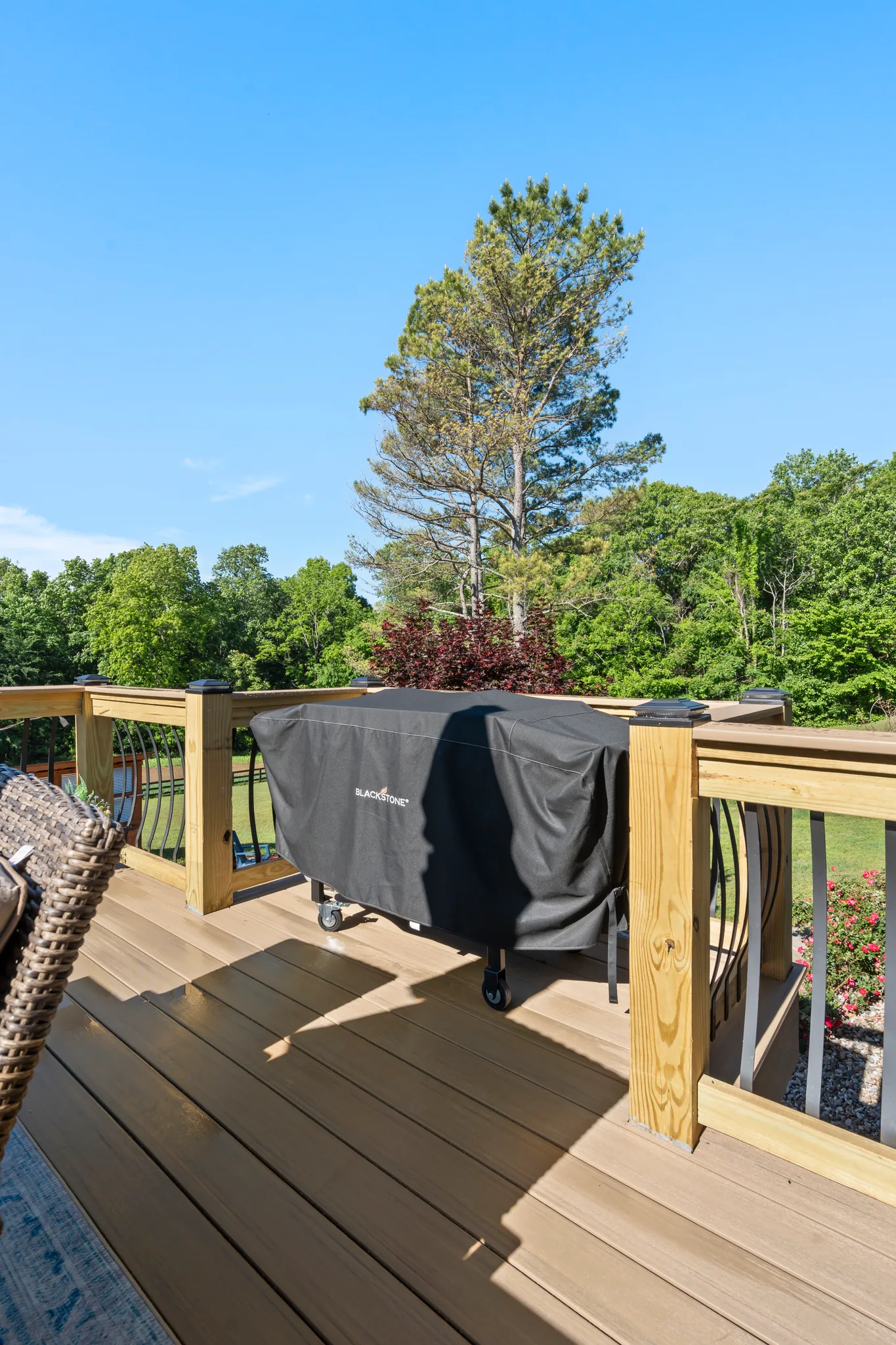 Composite deck with wood railing posts and curved metal balusters, holding a covered griddle on casters, with trees and lawn beyond