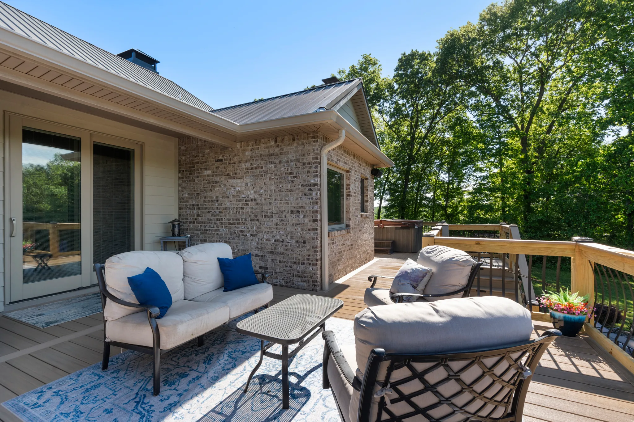 Composite deck with cushioned metal lounge furniture, blue patterned rug and glass table beside a brick house wall with sliding glass doors