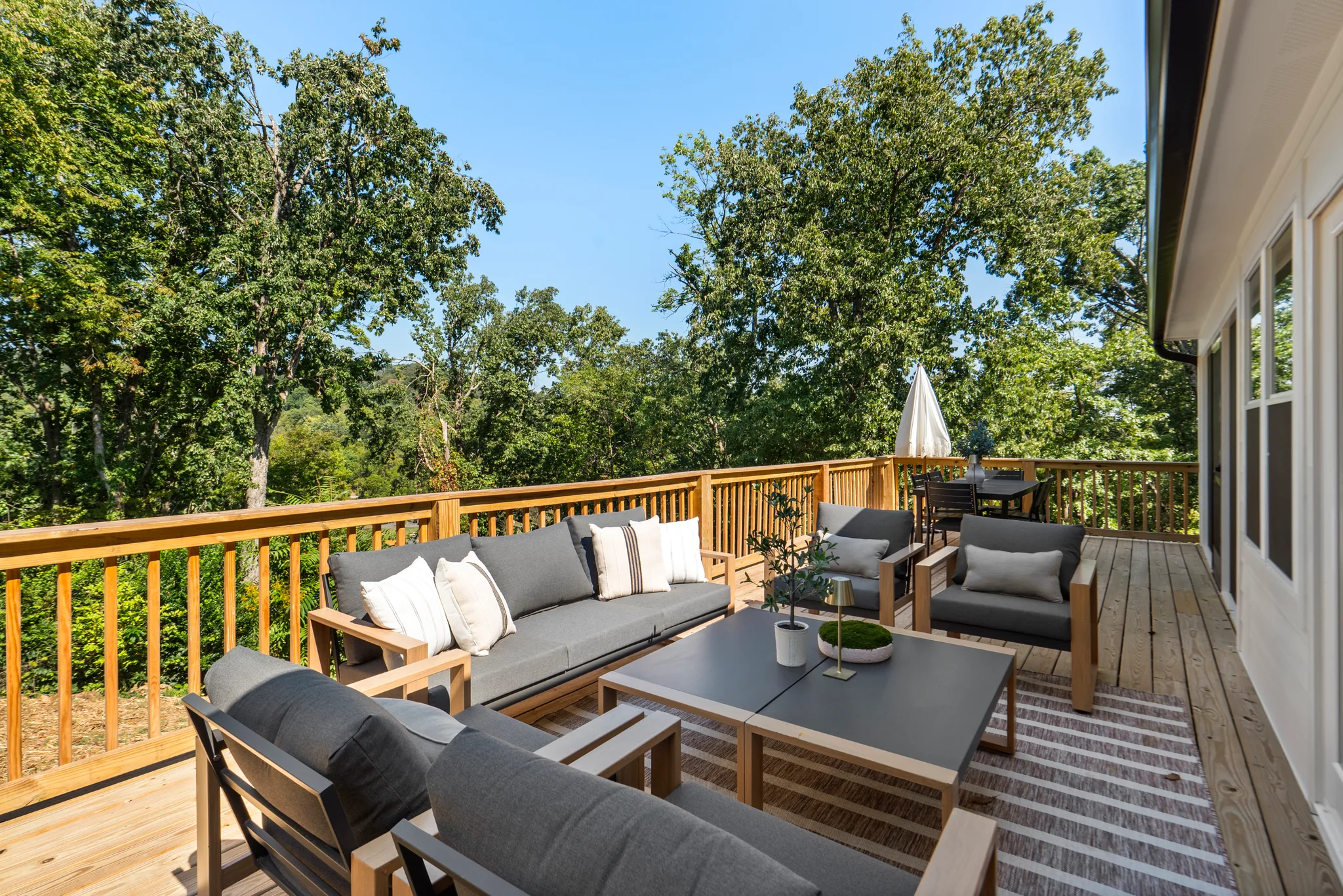 Wood deck with gray cushioned sofa and chairs, dark coffee table and striped rug, framed by natural wood railing and trees