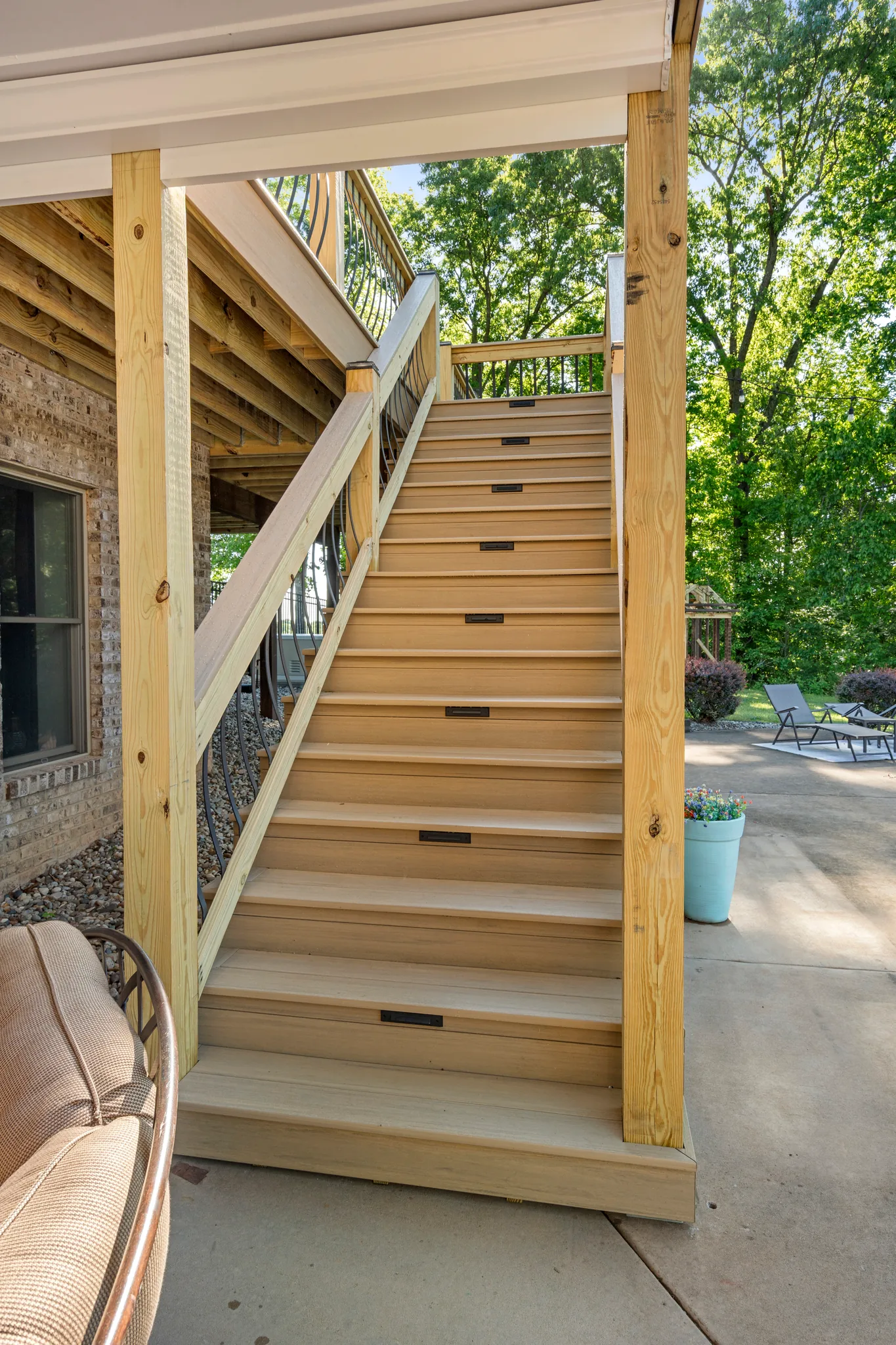 Wide wood deck stairs with recessed step lights descending to a concrete patio beside a brick home, framed by treated posts and trees