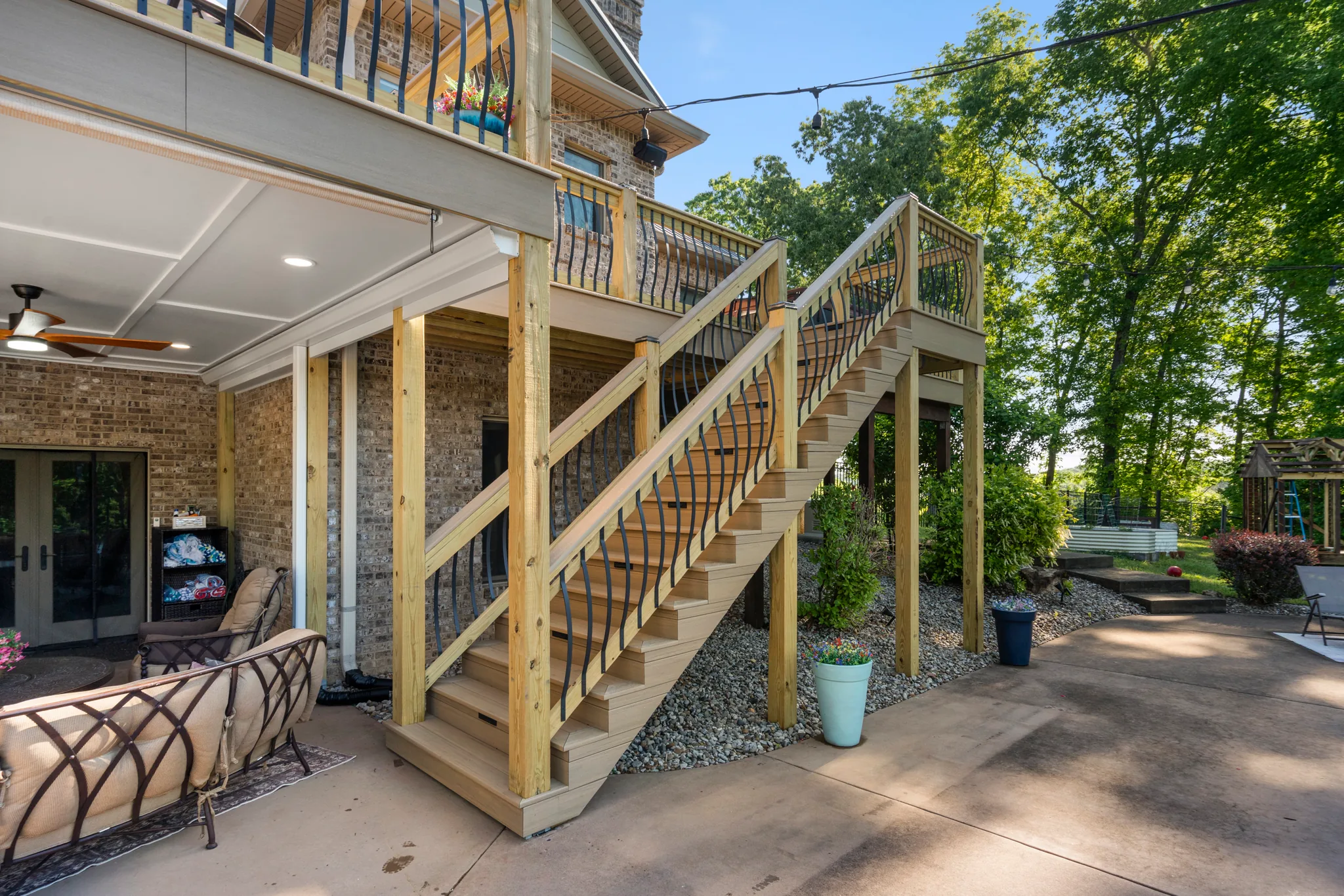 Wide wood deck staircase with branch-style metal balusters descending from an elevated deck to a concrete patio beside a brick home