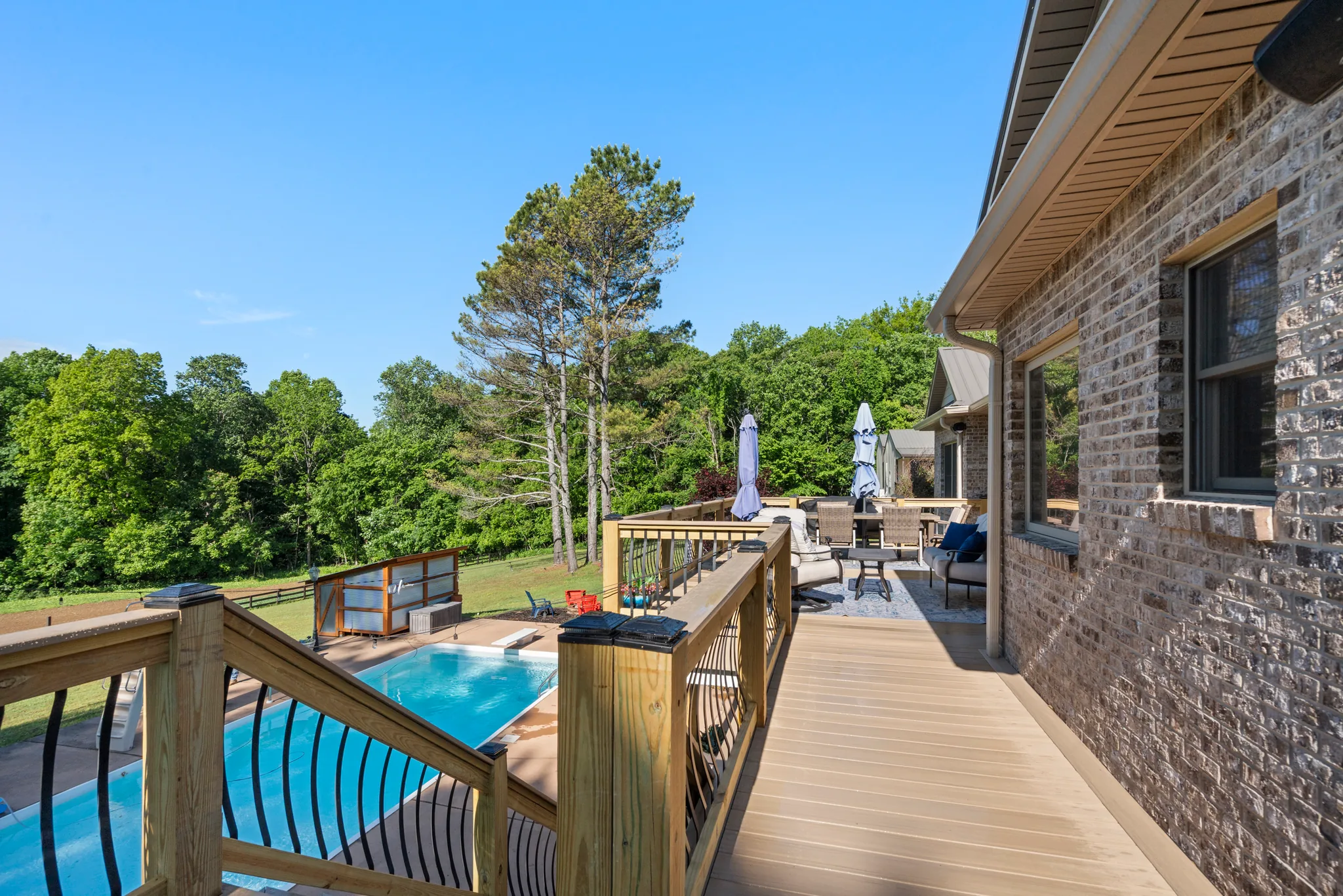 Composite deck walkway running beside a brick wall with a wood railing and dark balusters overlooking a rectangular pool and wooded yard