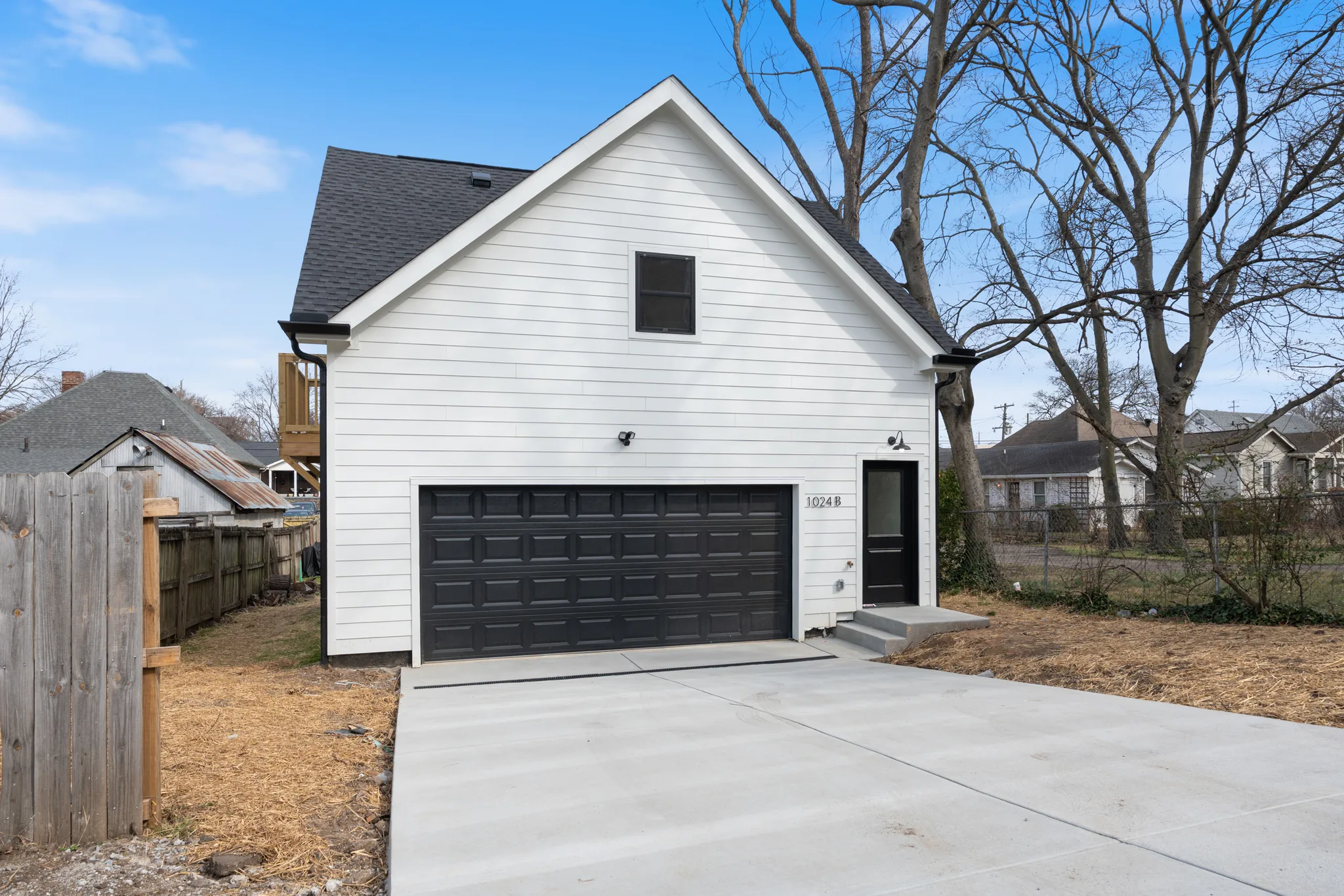 Detached two-car garage with white lap siding, black sectional door, dark shingled gable roof and a wide concrete driveway beside a wood fence