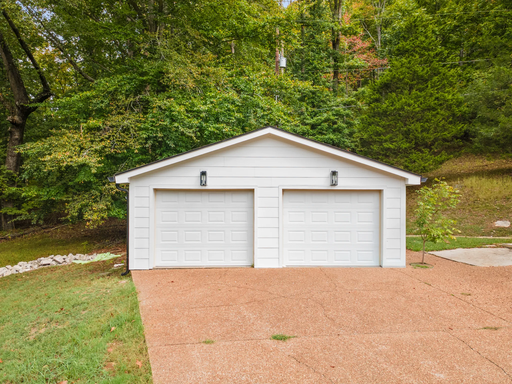 Detached two-car garage with white lap siding, two white paneled overhead doors, black sconces and an exposed aggregate driveway at a wooded slope