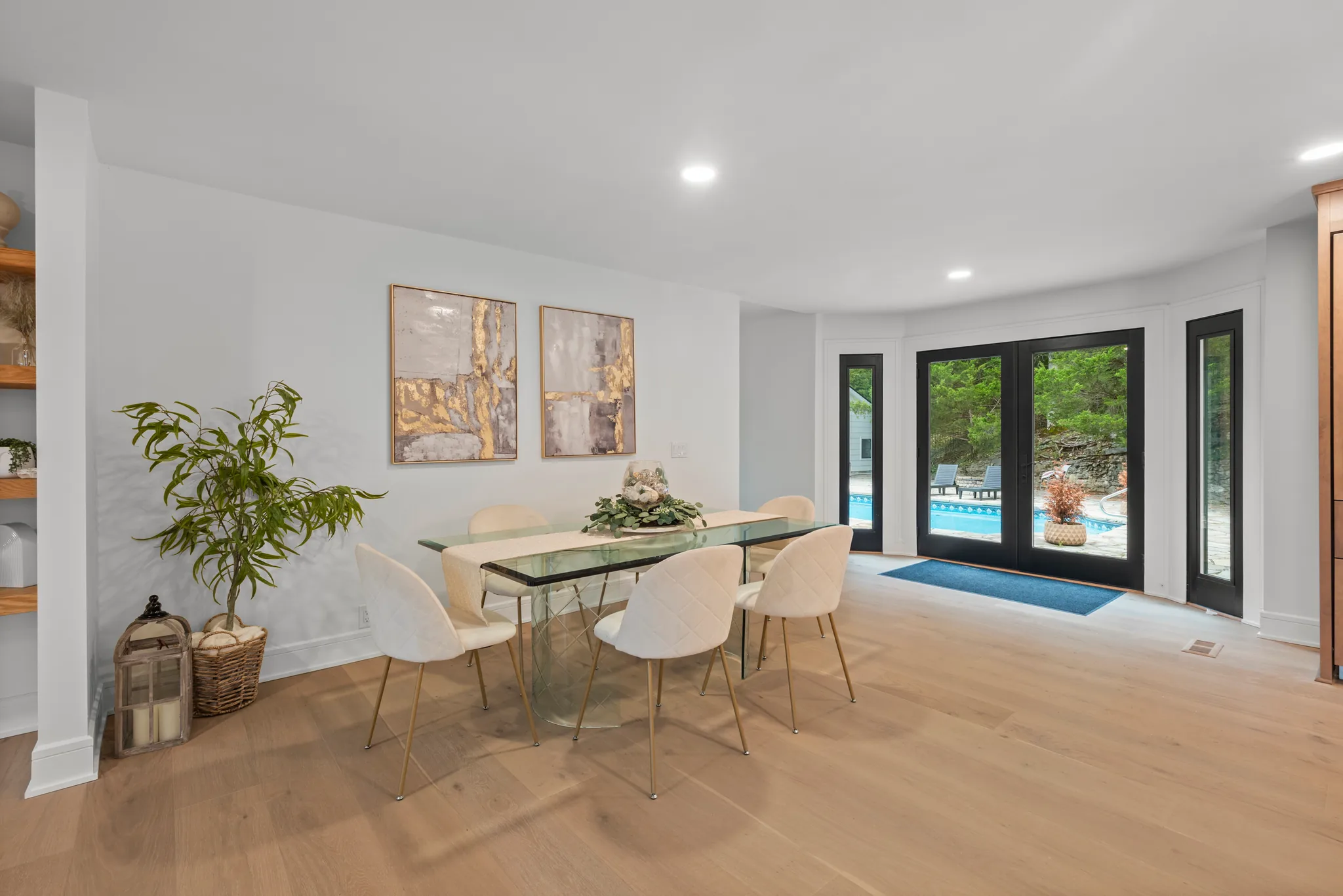 Dining area with glass table and white chairs on wide oak plank flooring, facing black French doors and sidelights overlooking a pool