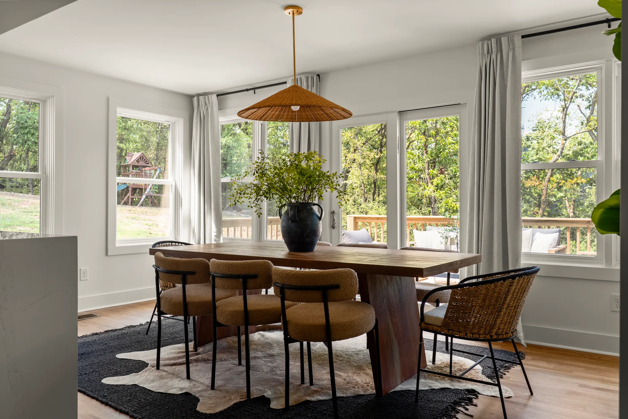 Dining area with a walnut trestle table, upholstered chairs and a woven pendant light beside windows and glass doors overlooking a deck and trees