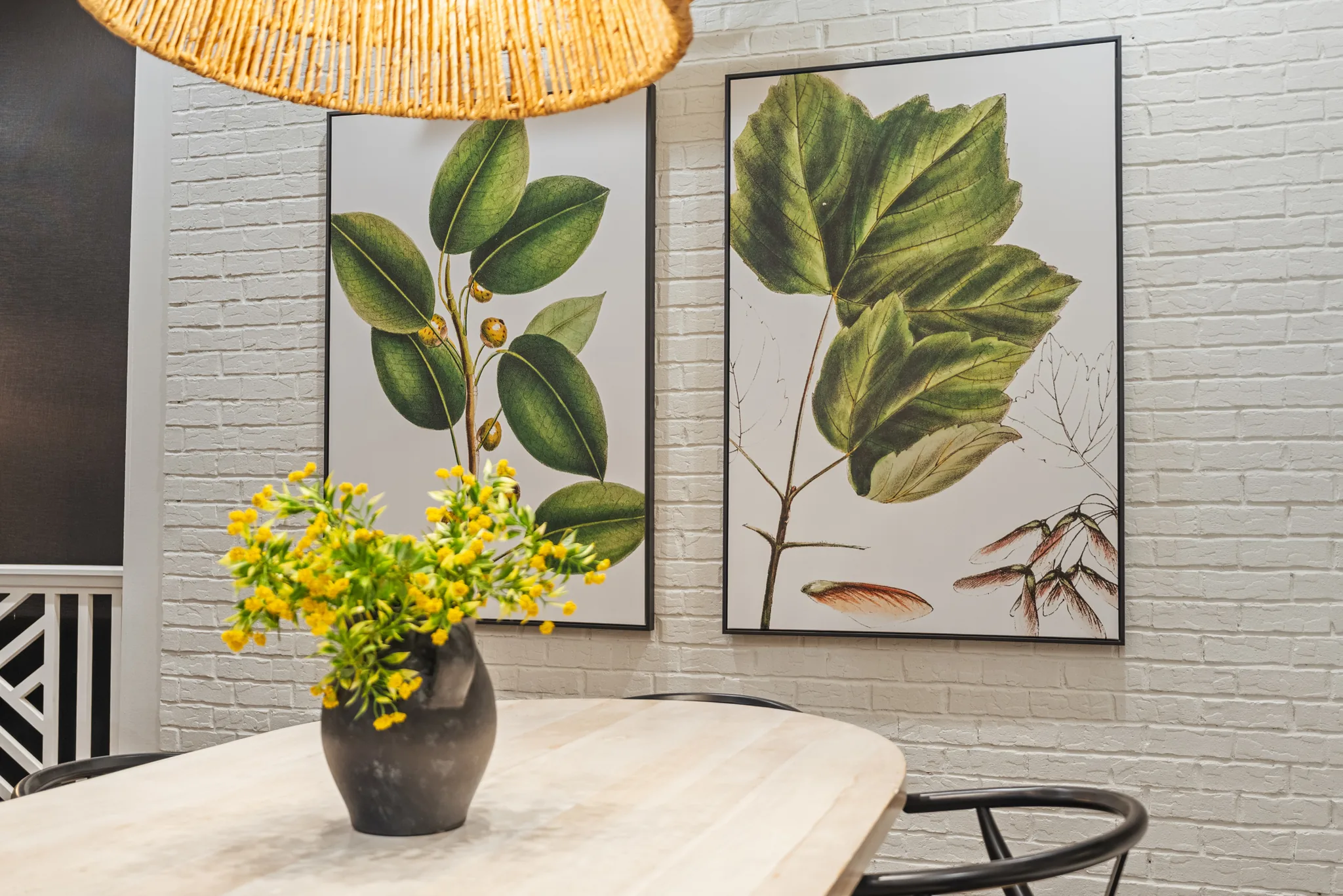 Dining table with a woven pendant shade beside a painted white brick wall hung with two framed botanical prints and black chairs