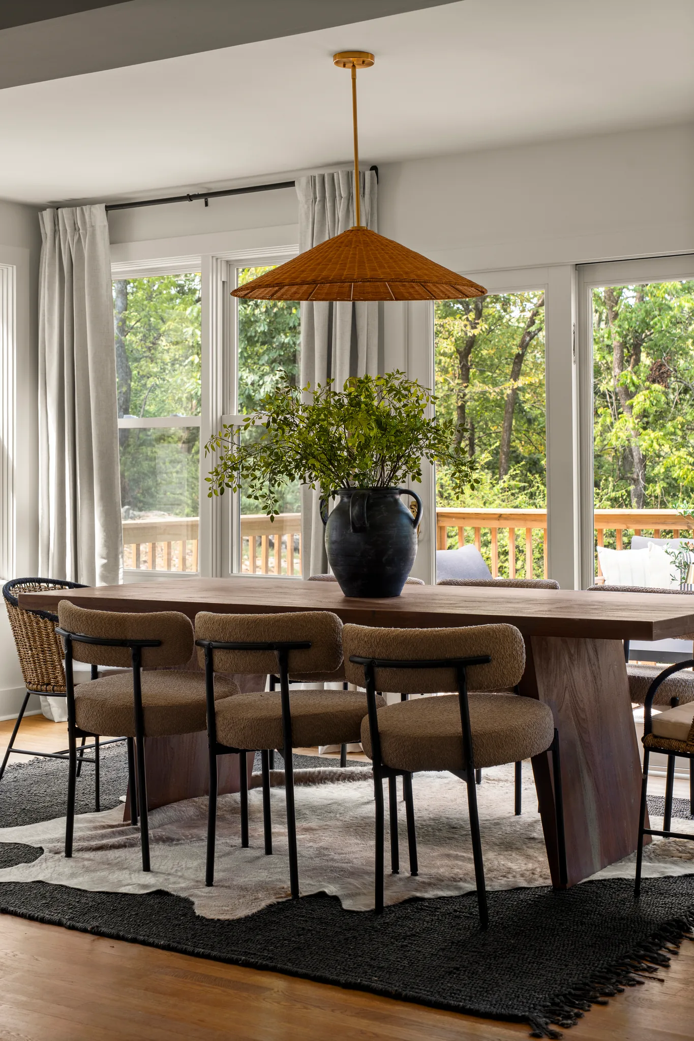 Dining room with a walnut table, boucle chairs and a woven pendant light in front of windows overlooking a wood deck and trees