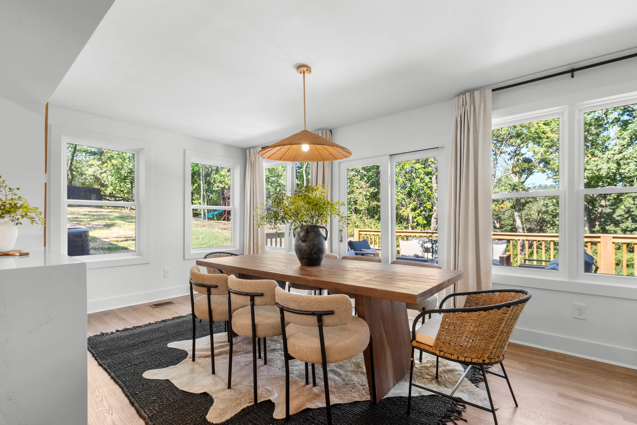 Dining area with wood table, upholstered chairs and woven pendant, surrounded by white-trimmed windows overlooking a wood deck and trees