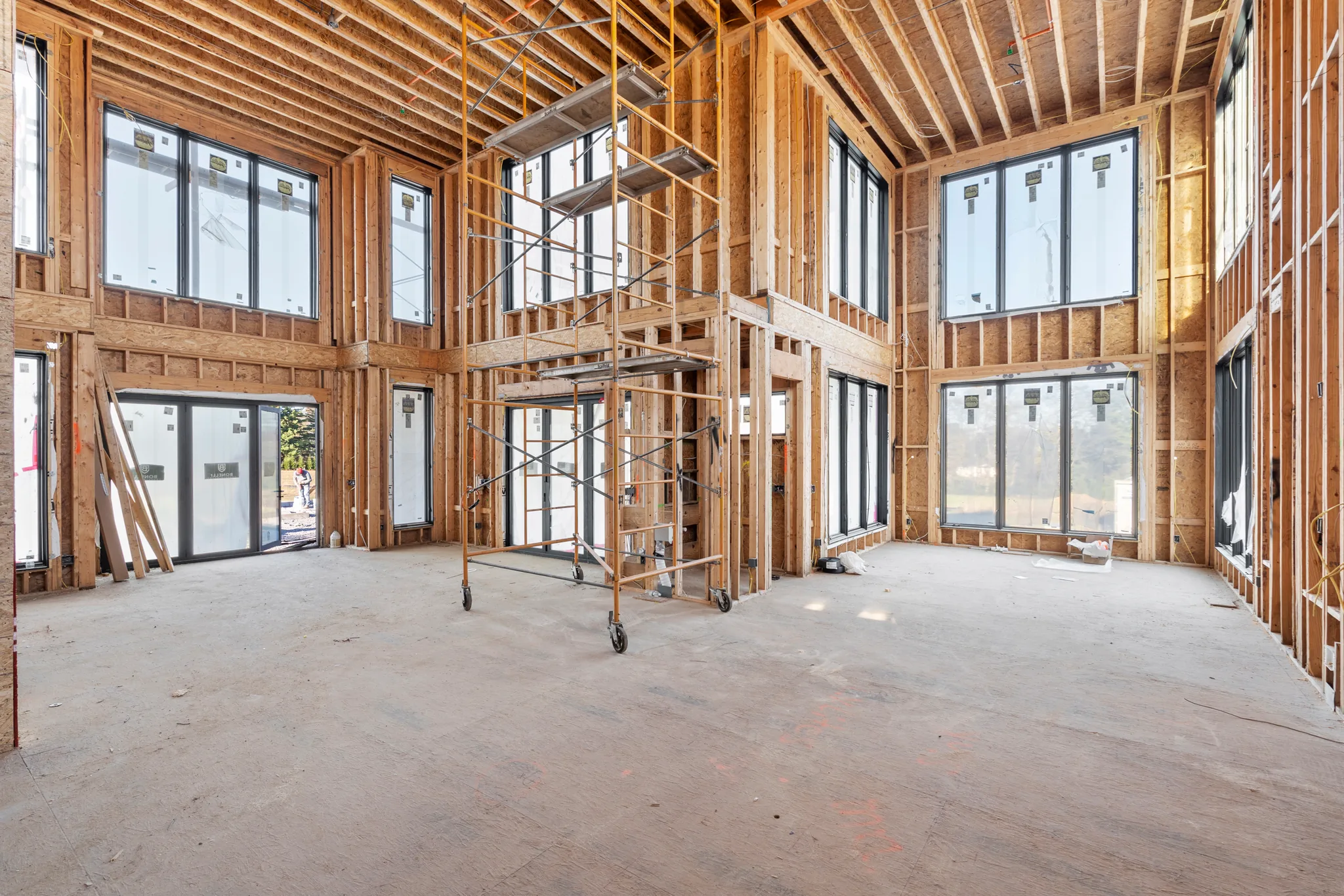 Two-story room under construction with wood stud framing, installed black windows and sliding doors, and a rolling scaffold tower on plywood flooring