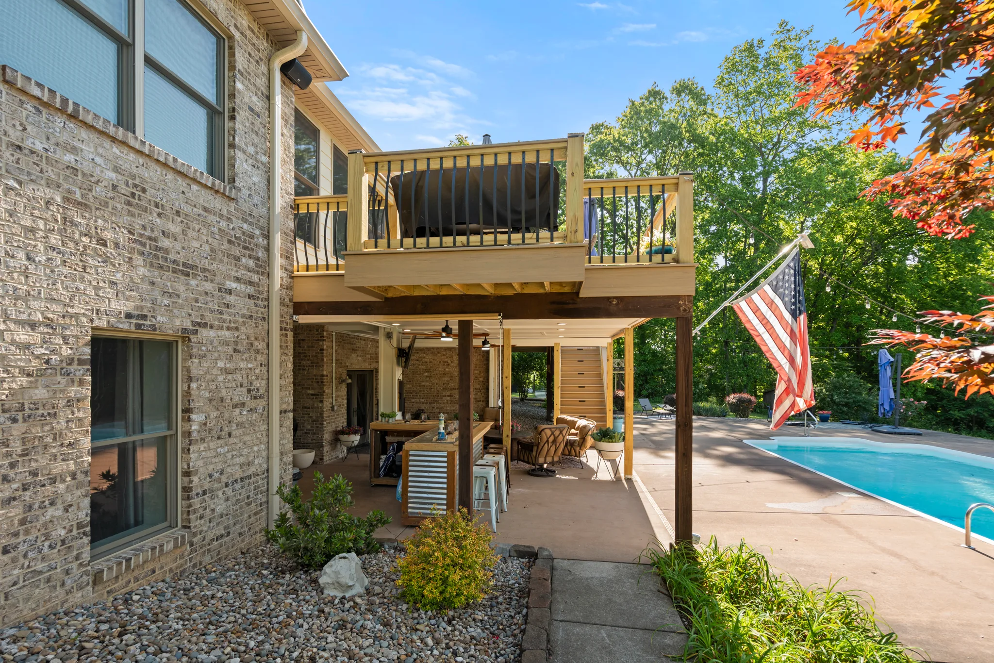 Second-story wood deck with spindle railing built over a covered patio with a bar, stools and ceiling fans beside an in-ground pool