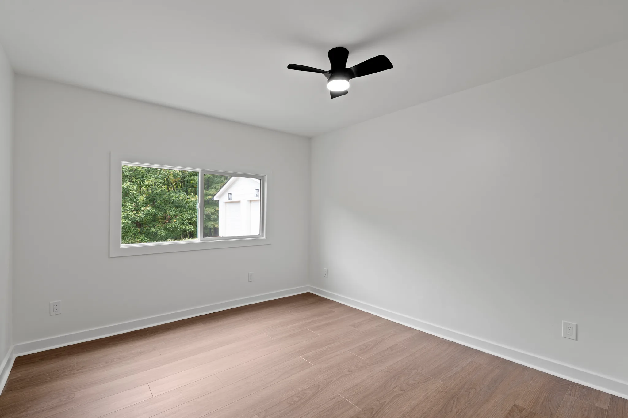 Empty bedroom with light gray walls, wood-look plank flooring, a sliding window over trees, and a black three-blade ceiling fan with light